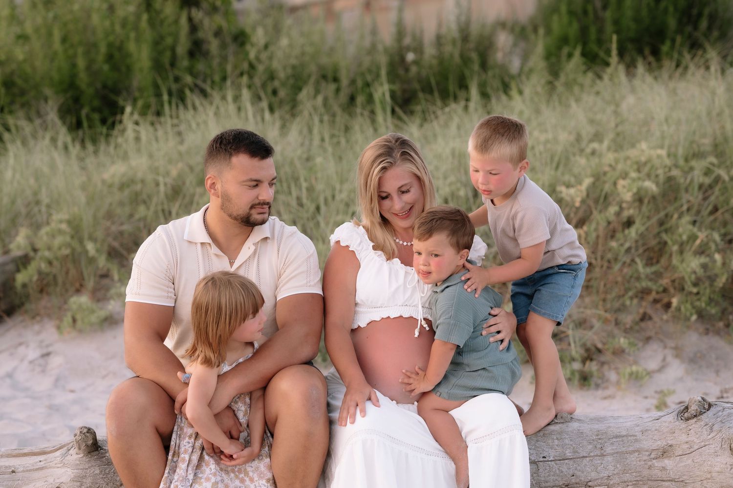 Pregnant woman sits on beach with husband and three young children surrounded by green dunes in a family maternity photo.
