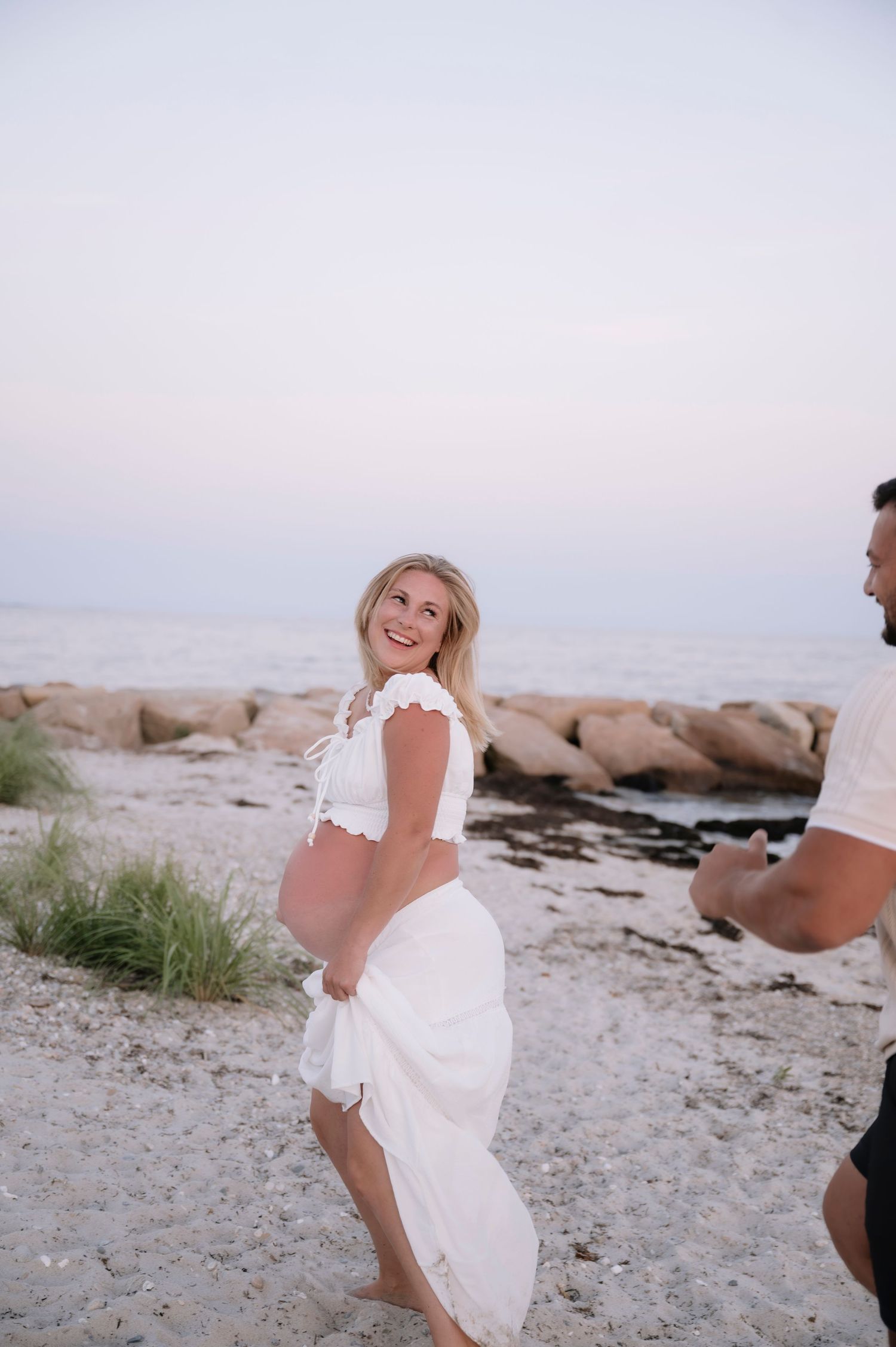 Smiling pregnant woman in white lace dress twirling on beach at dusk with partner nearby.