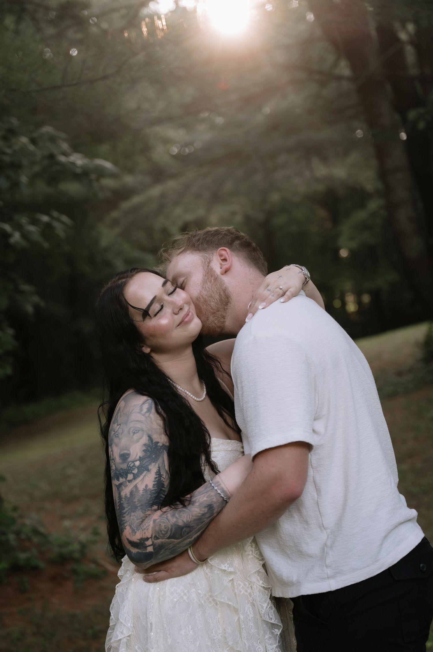 Couple embracing in forest at golden hour, woman with sleeve tattoos in white dress, man kissing her cheek.