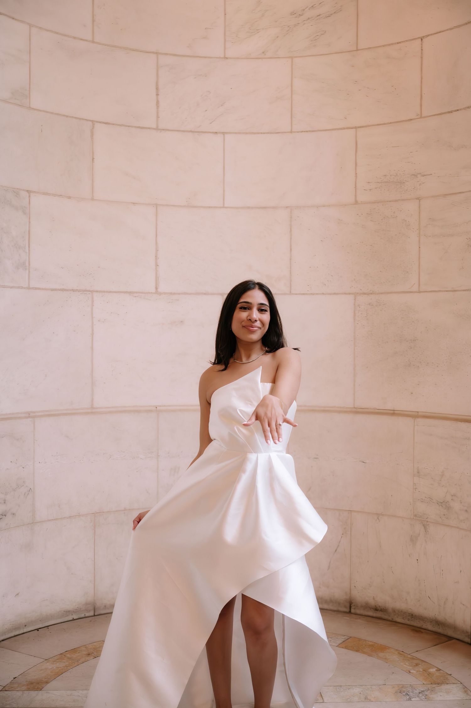 A person in an elegant white wedding dress poses gracefully against a curved marble wall in a romantic architectural setting.