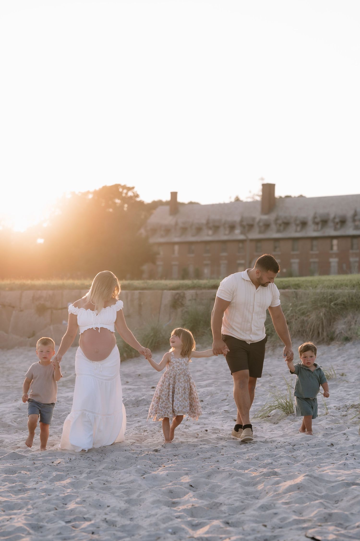 Family of five walking on sandy beach at golden sunset, pregnant mother in white dress holding hands with children.