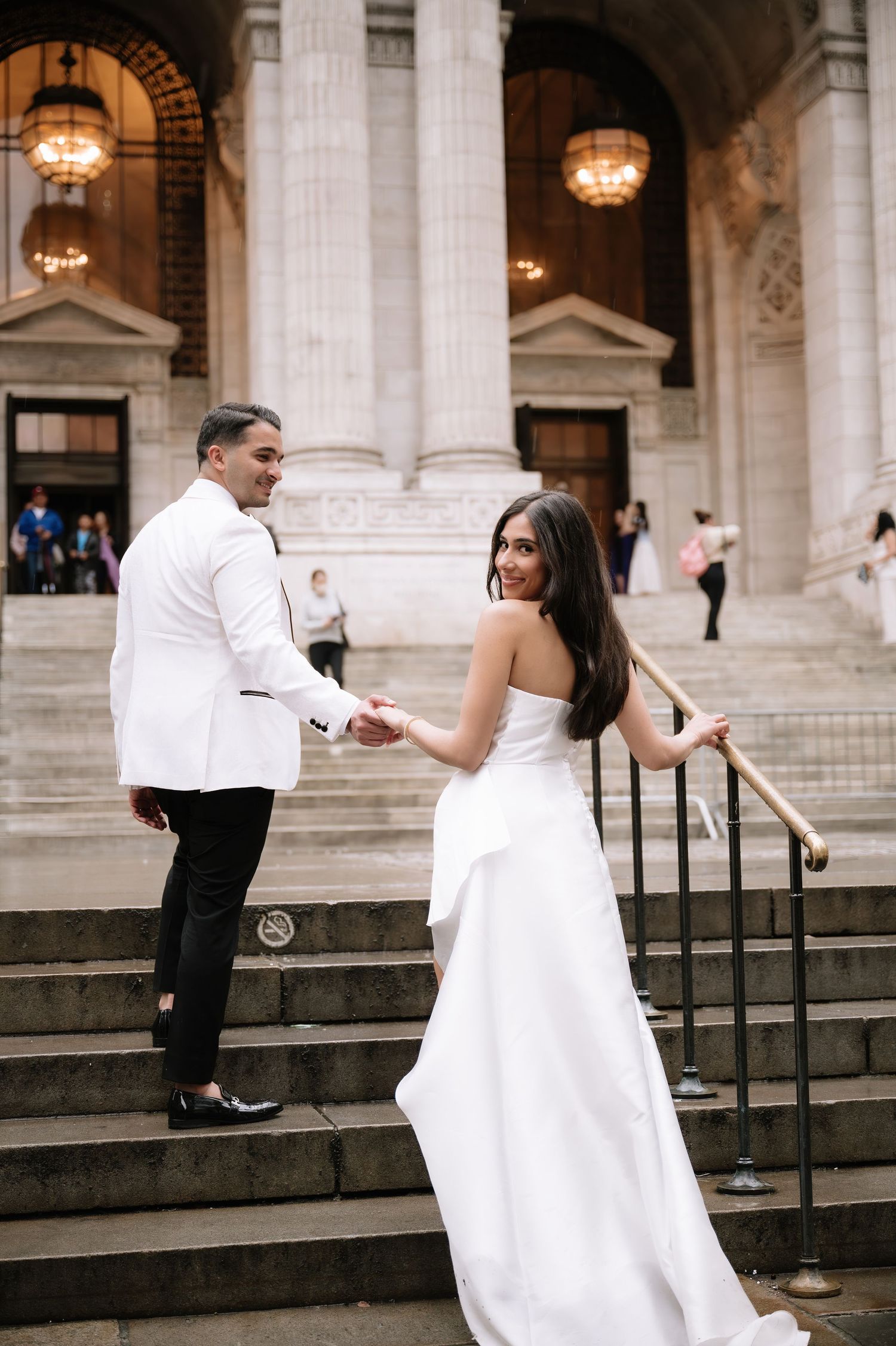A couple in white wedding attire pose on the grand steps of a historic building with ornate columns and lighting fixtures.