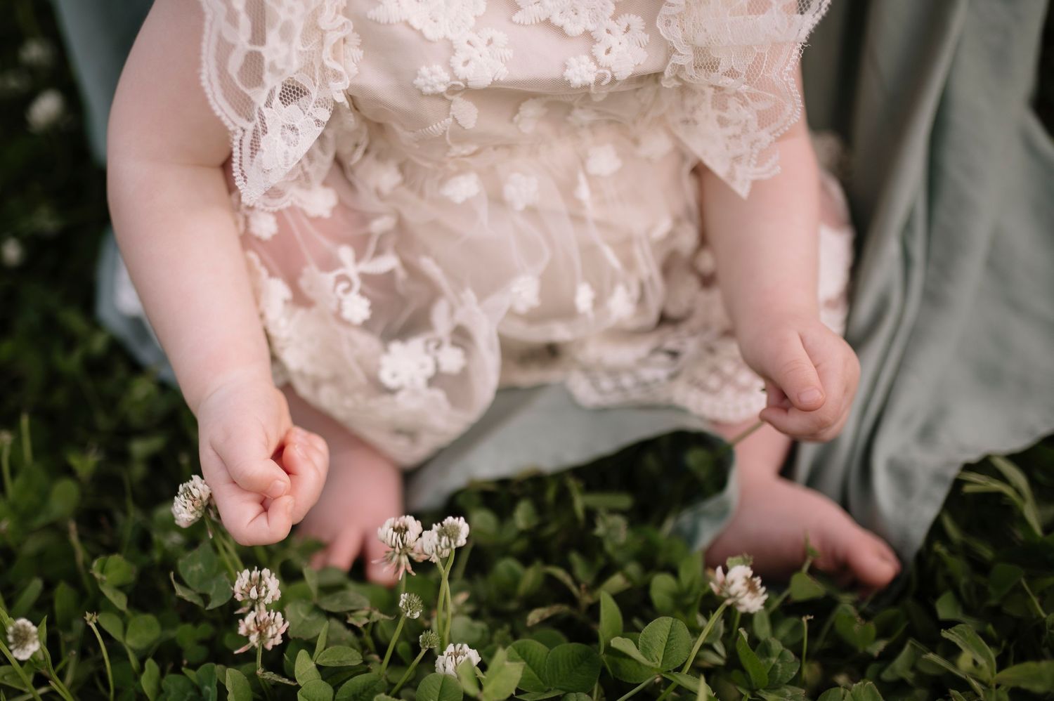 Tiny bare feet in a white lacy dress reaching down to touch white clover flowers in green grass.
