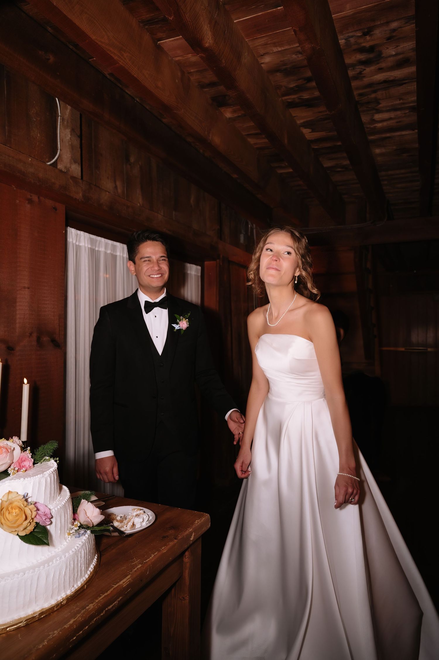 Bride in white strapless gown and groom in black tuxedo stand together near wedding cake in rustic wood-paneled venue.