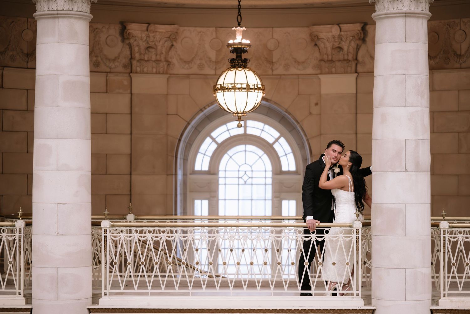 Bride and groom embrace on elegant balcony with chandelier and arched windows in grand wedding venue.