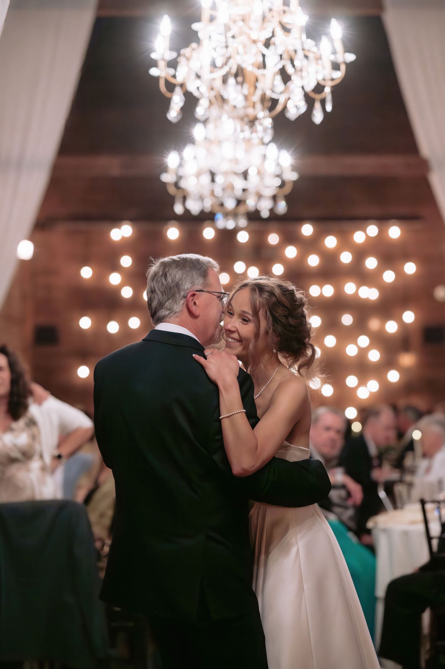 Bride shares a joyful father-daughter dance under a crystal chandelier at her wedding reception.