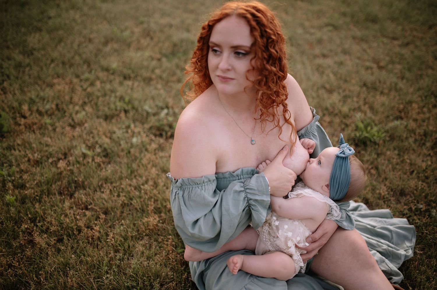 A woman with curly red hair in an off-shoulder blue dress poses on grassy field at sunset.