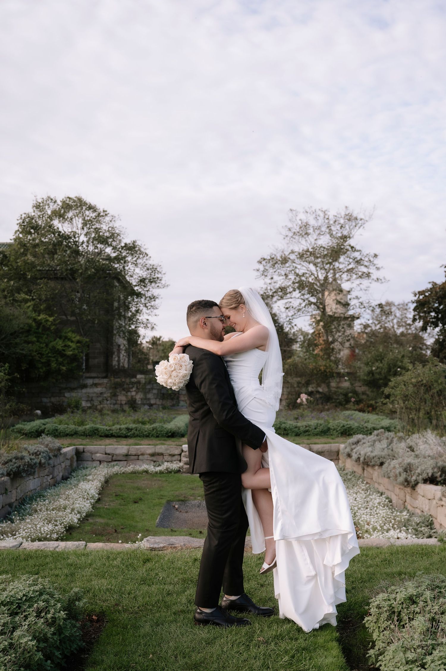 A romantic wedding couple shares an intimate moment in a garden setting with stone walls and trees.