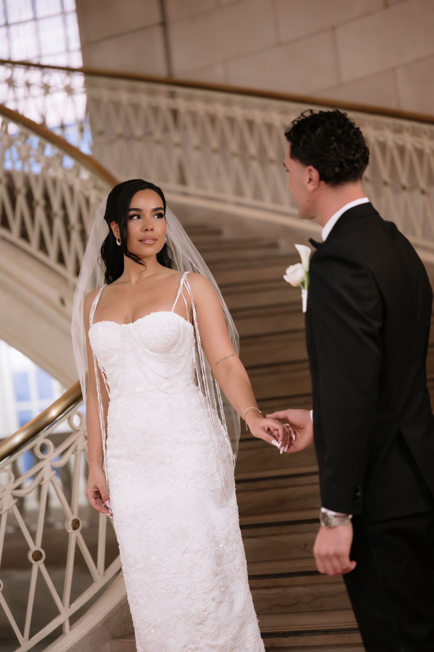Bride in lace gown and veil holds groom's hand on ornate staircase.
