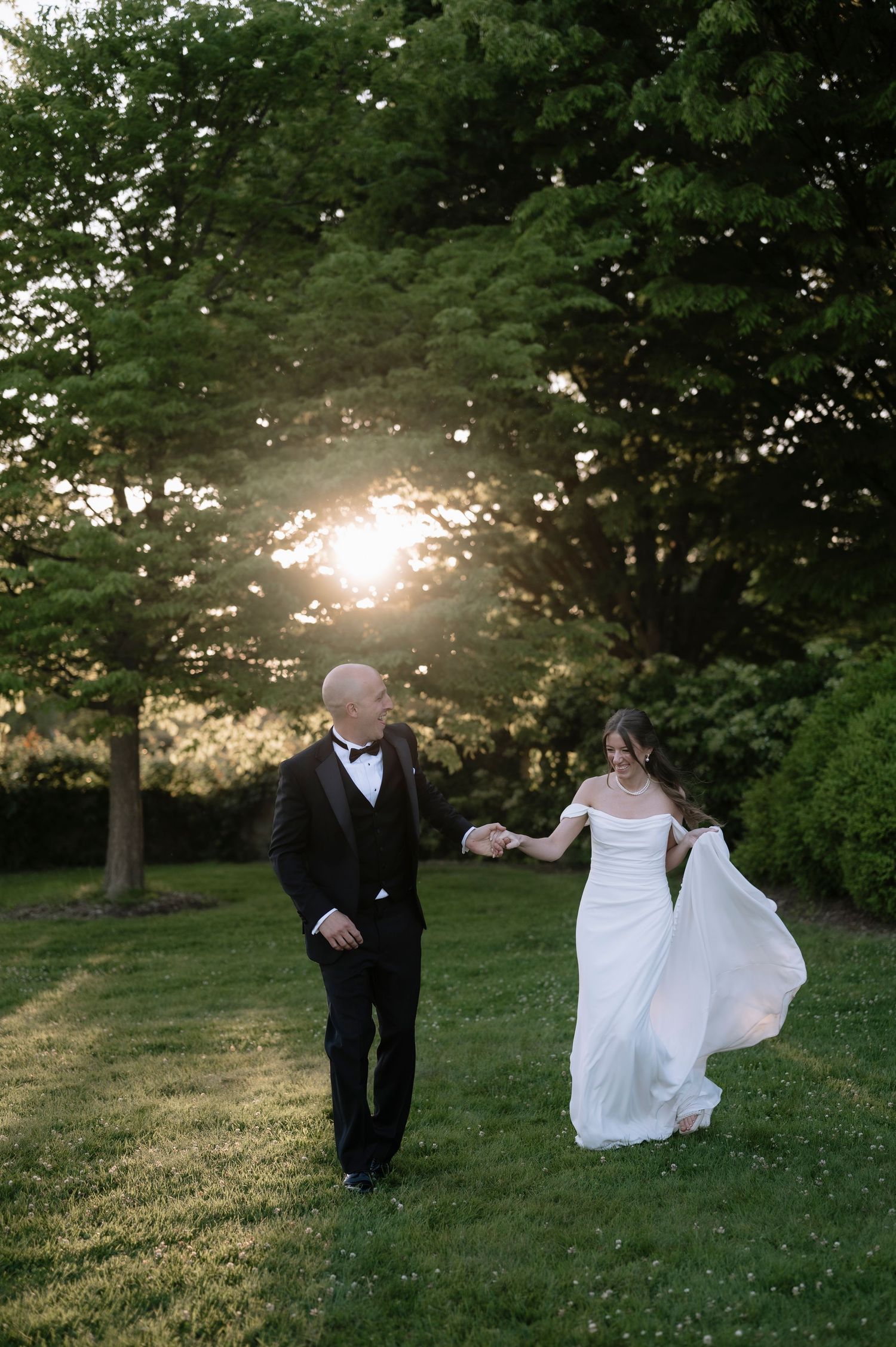 A couple in formal wedding attire walking together on a grassy lawn at sunset with trees in the background.
