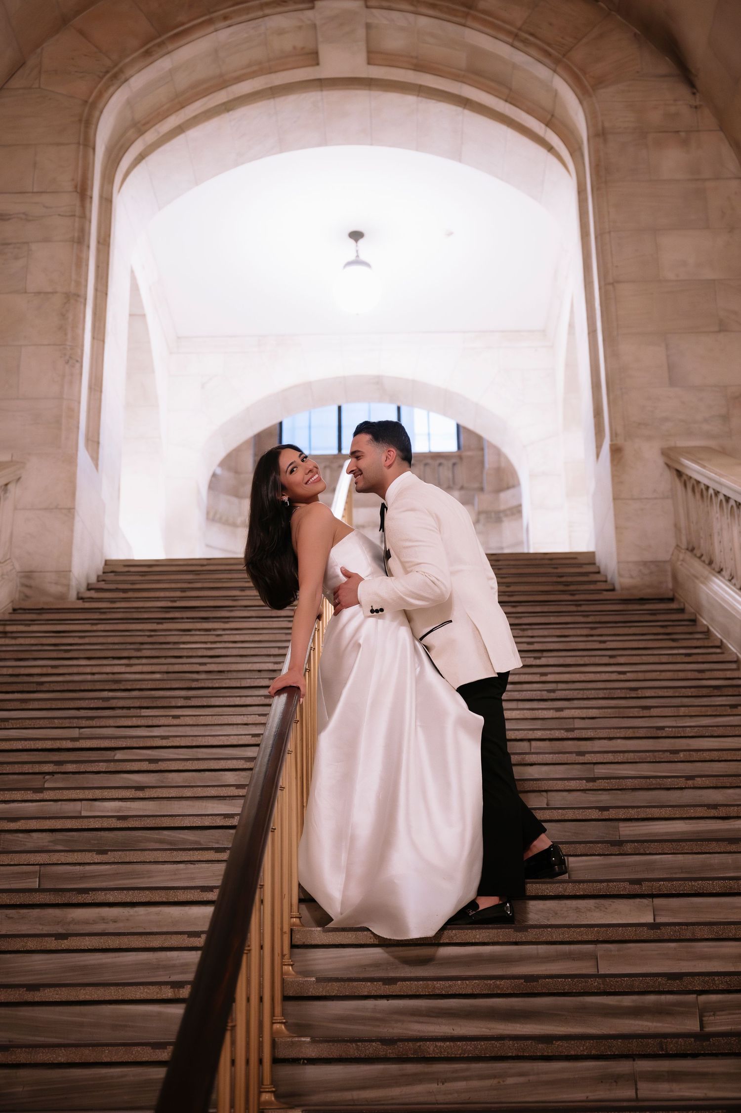 Couple in wedding attire sharing romantic moment on grand marble staircase.
