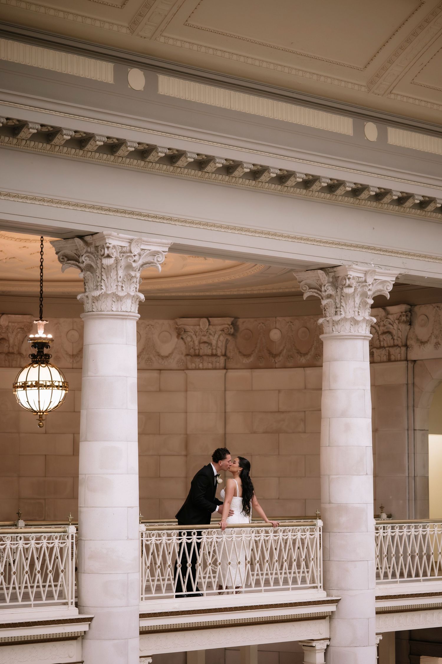 Bride and groom share a romantic kiss on an elegant balcony with ornate white columns in a grand historic venue.
