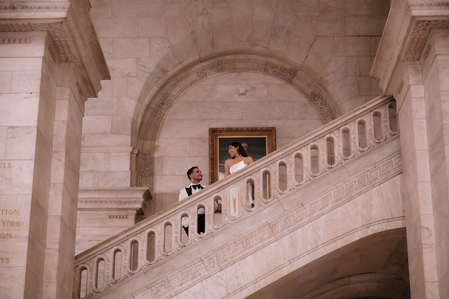 A couple stands on an elegant marble staircase in a classical building interior with arched architecture.