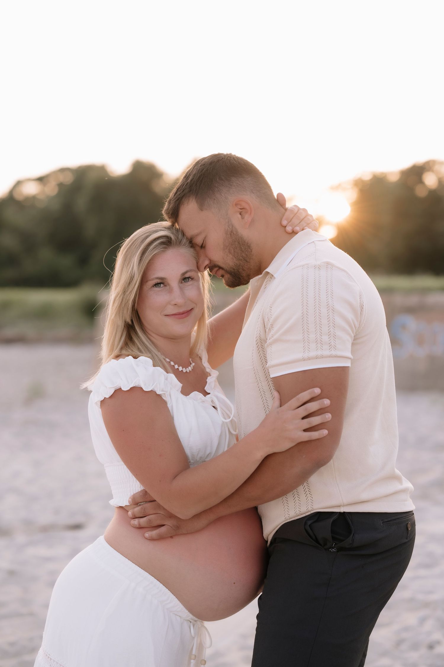 Pregnant couple embracing on a sandy beach at golden sunset, woman in white dress with pearl necklace, man in cream shirt.