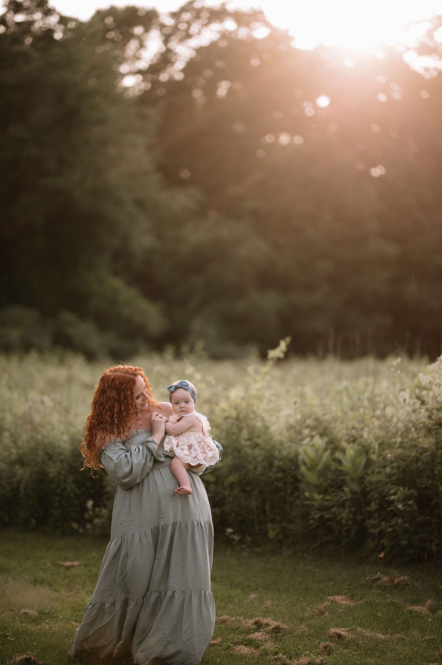 A woman in a flowing gray dress holds a baby in a sunlit meadow during golden hour.