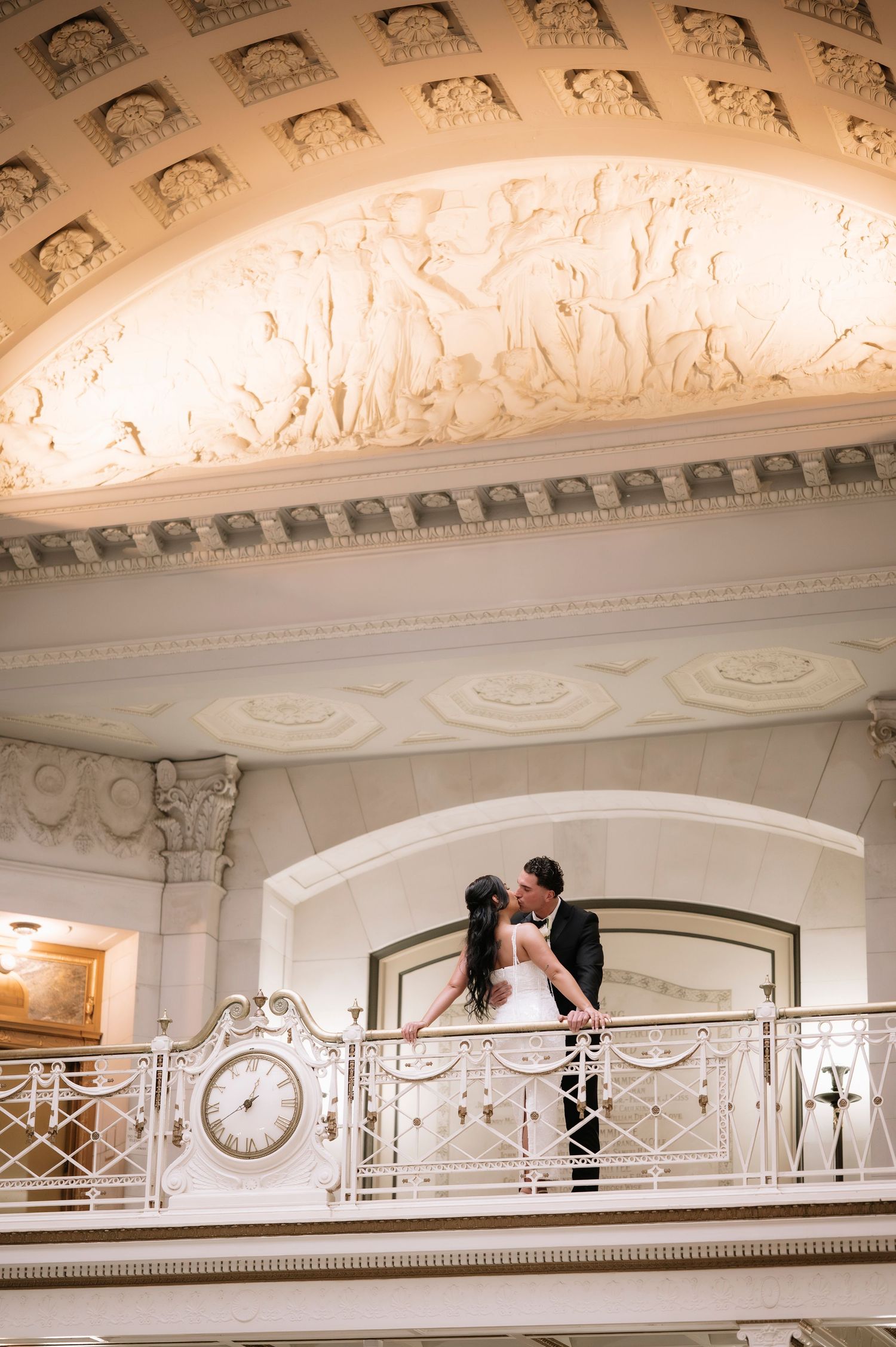 Bride and groom share a kiss on an ornate balcony with decorative ceiling reliefs at a grand wedding venue.