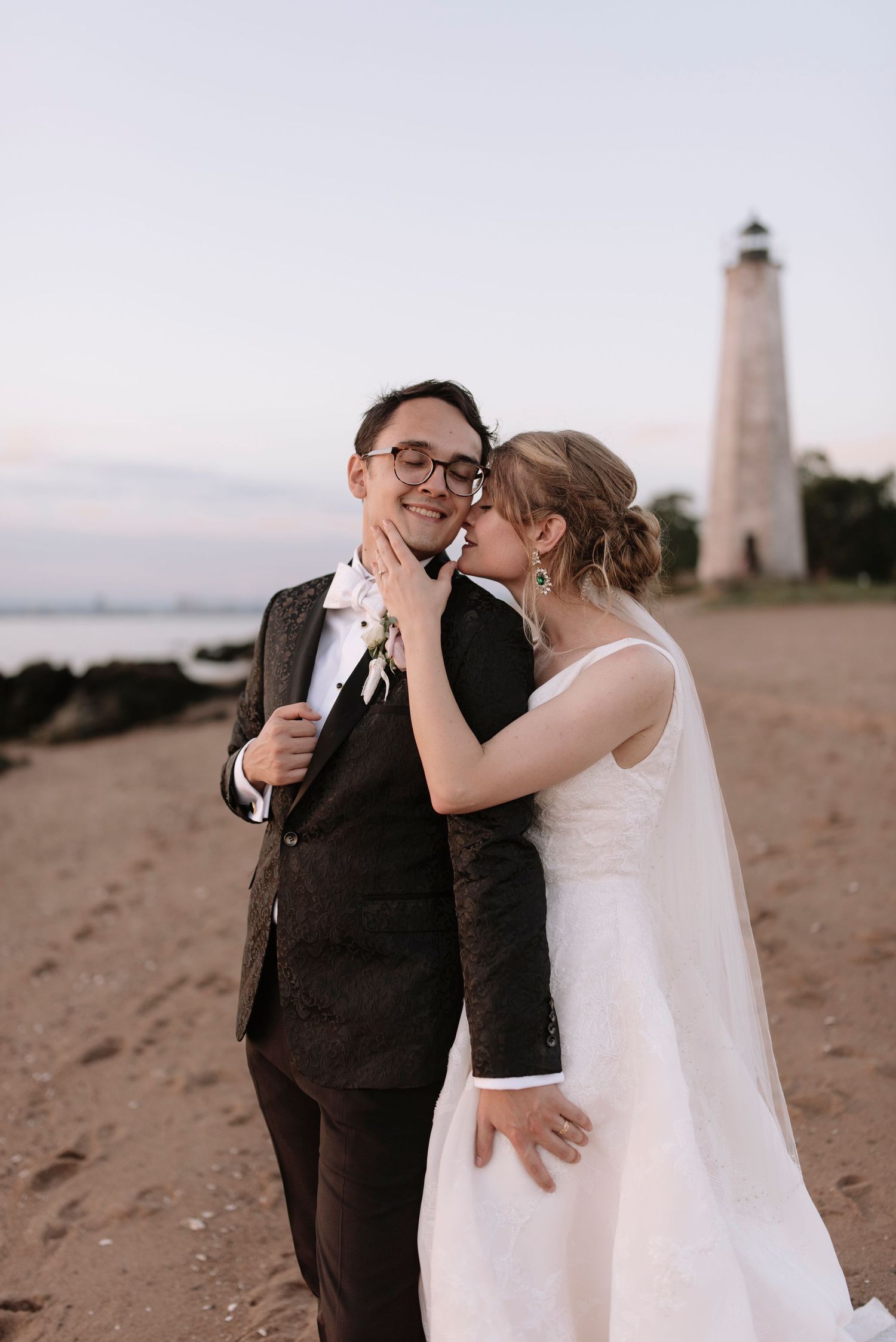 Romantic couple embracing at sunset near lighthouse on sandy beach.