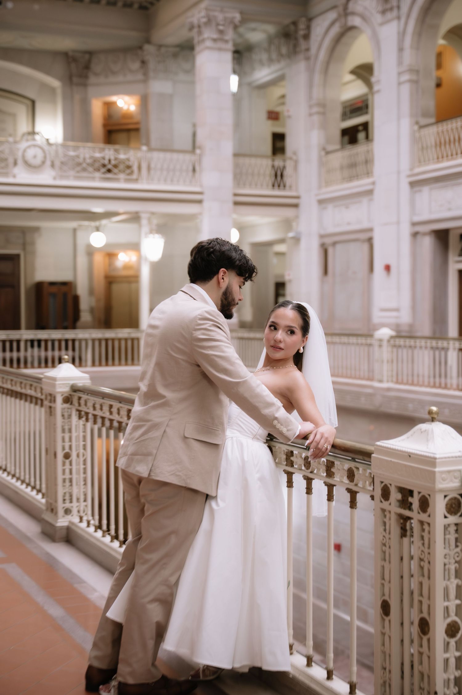 A couple in wedding attire share romantic moments on an elegant white marble staircase in a grand historic building.