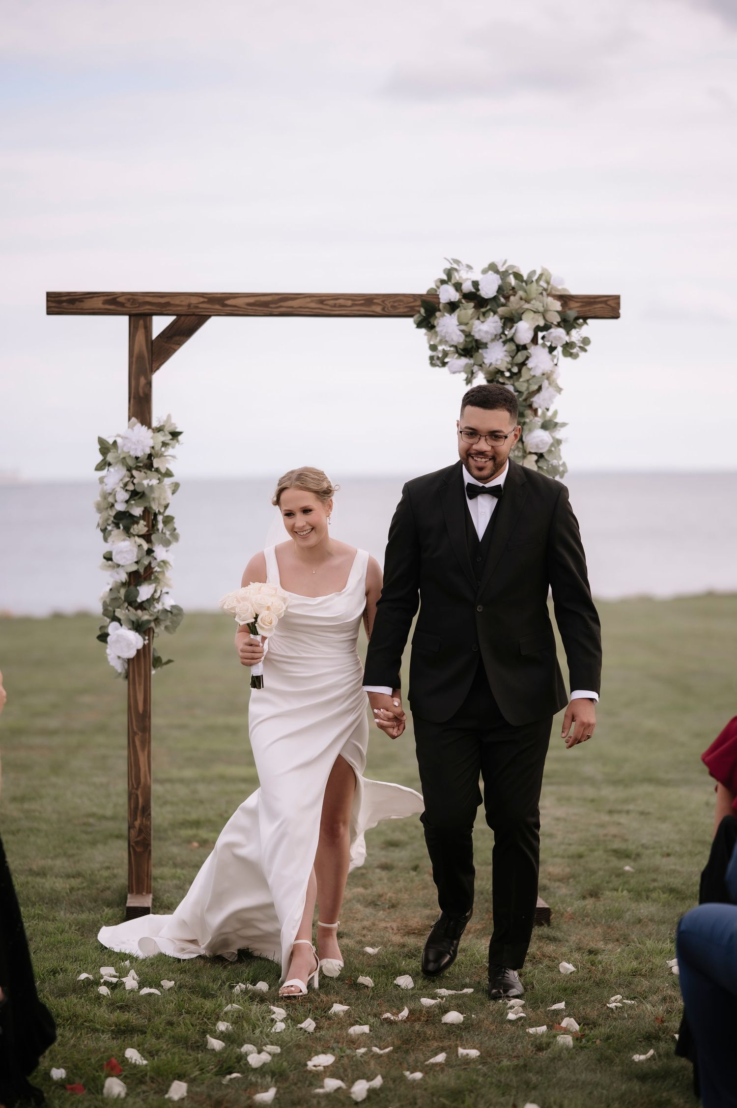 Newlyweds celebrate joyfully under a rustic wooden arch decorated with white flowers in an outdoor ceremony.