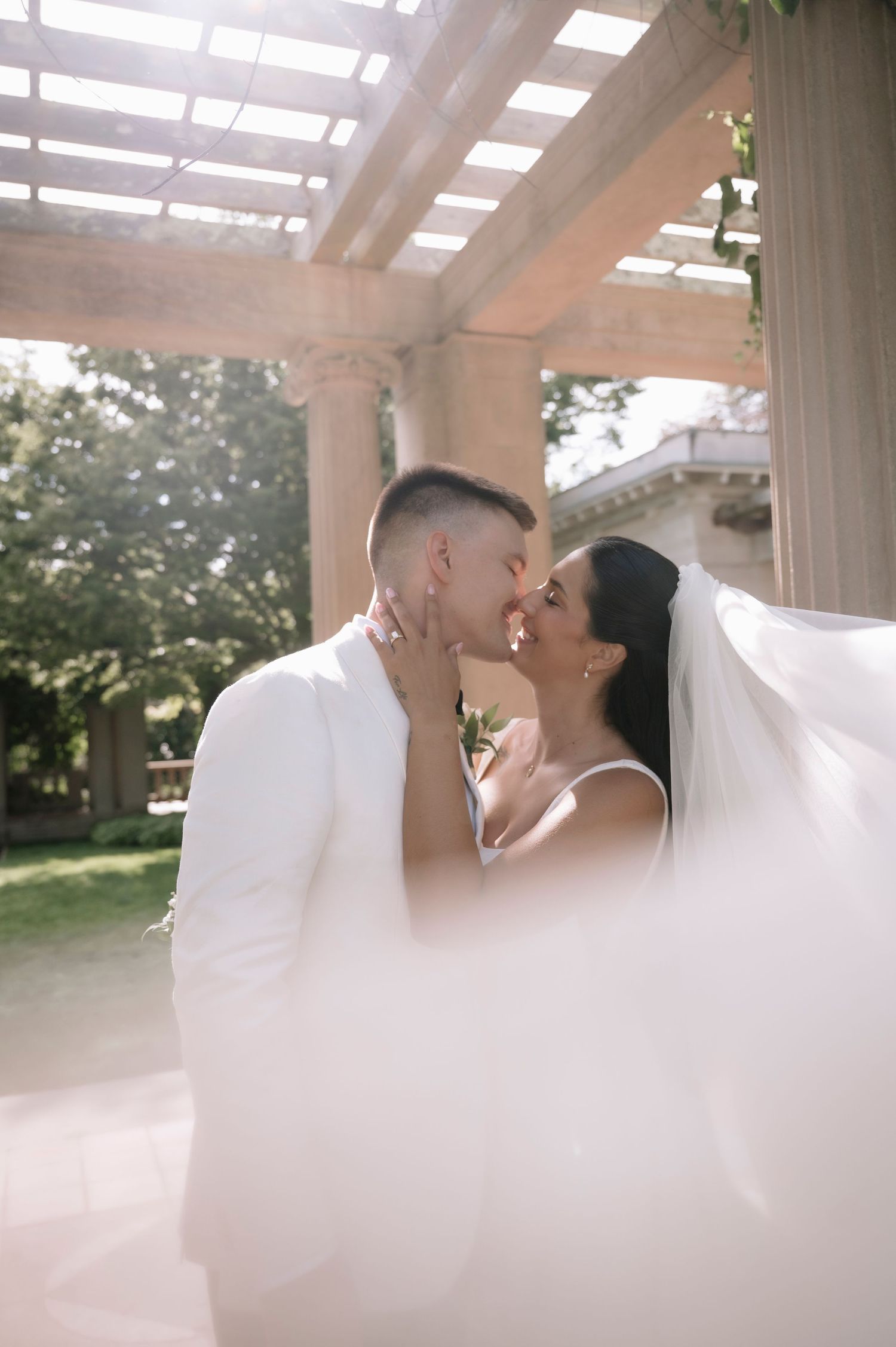 Bride and groom kiss tenderly under a pergola as the wedding veil billows in the breeze.