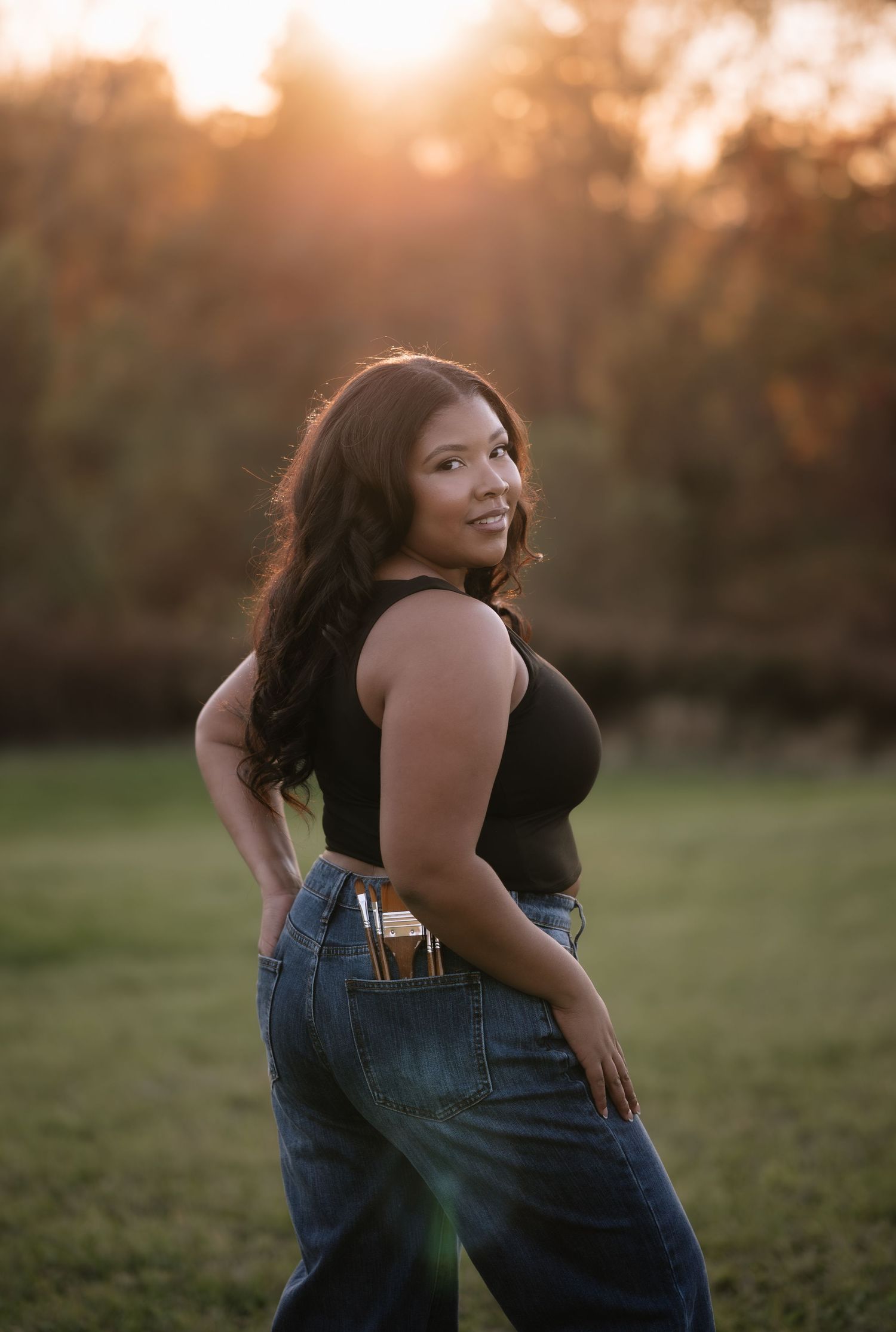 A woman in a black top and jeans poses outdoors during golden hour sunset in a grassy field.