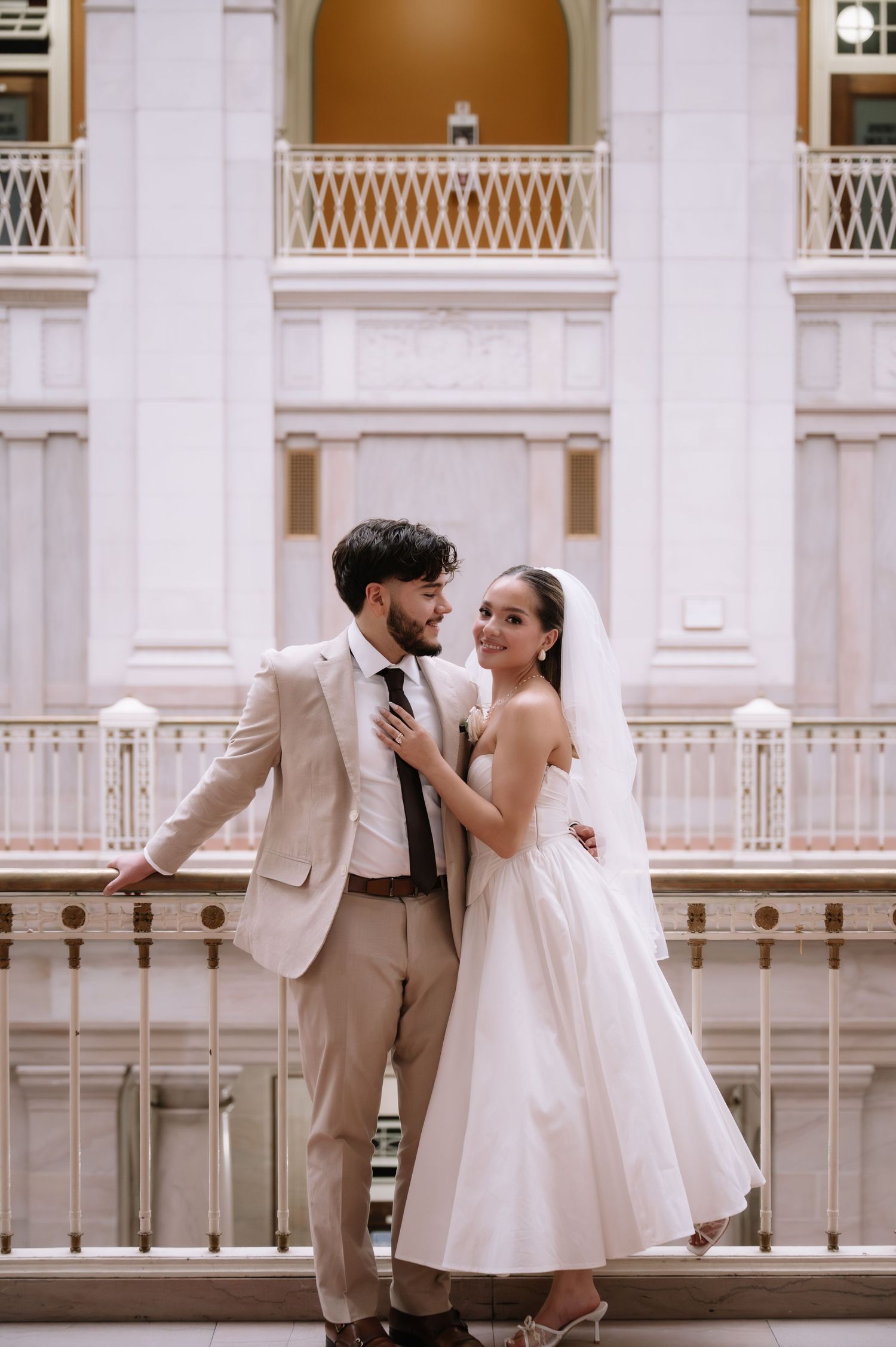 A couple in wedding attire pose together on an elegant white balcony with classical architectural details.