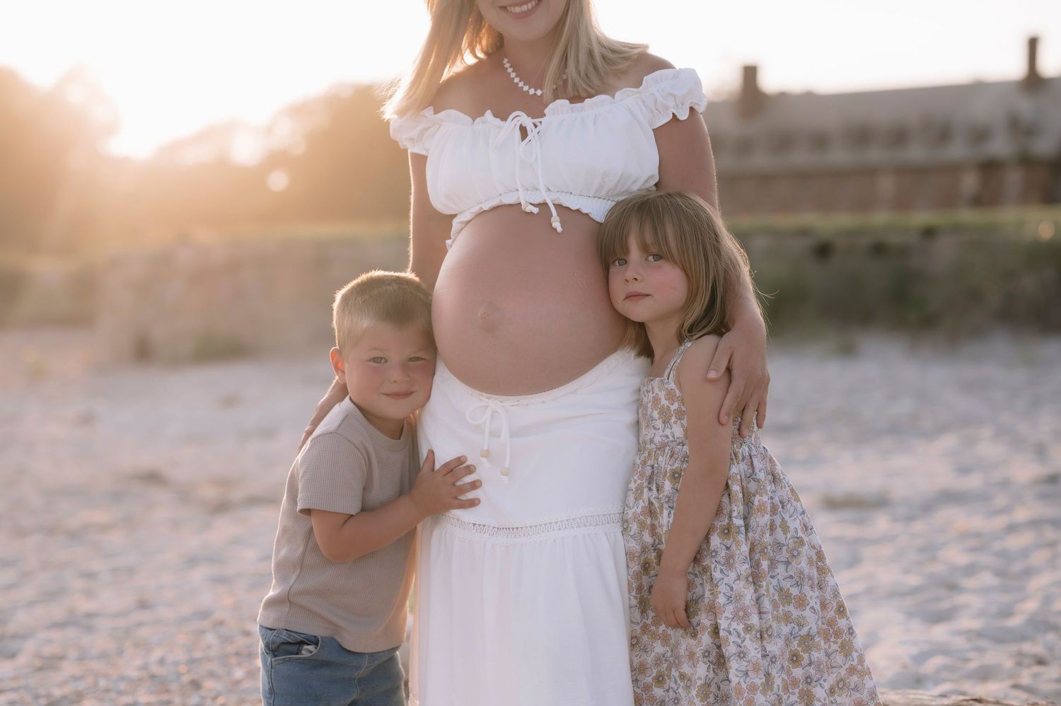 Pregnant woman in white dress poses with two children on a sunny beach during golden hour maternity photoshoot.