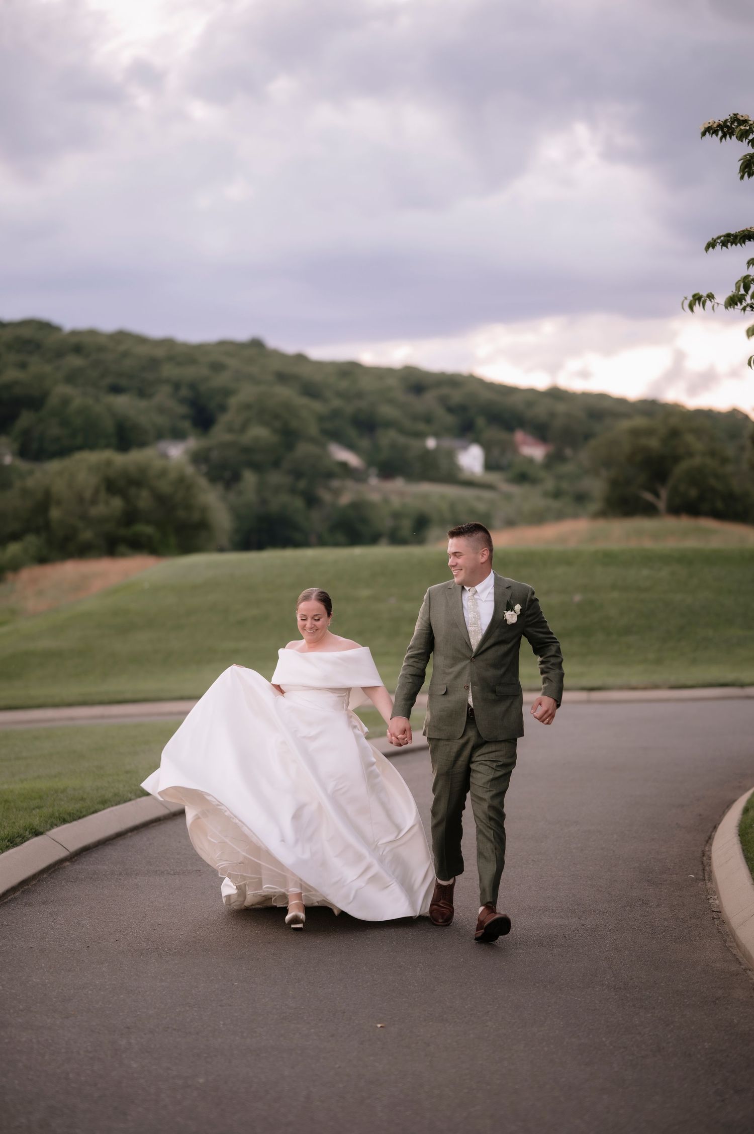 A bride and groom walk hand-in-hand on a path, her white gown flowing, as green hills stretch behind them.