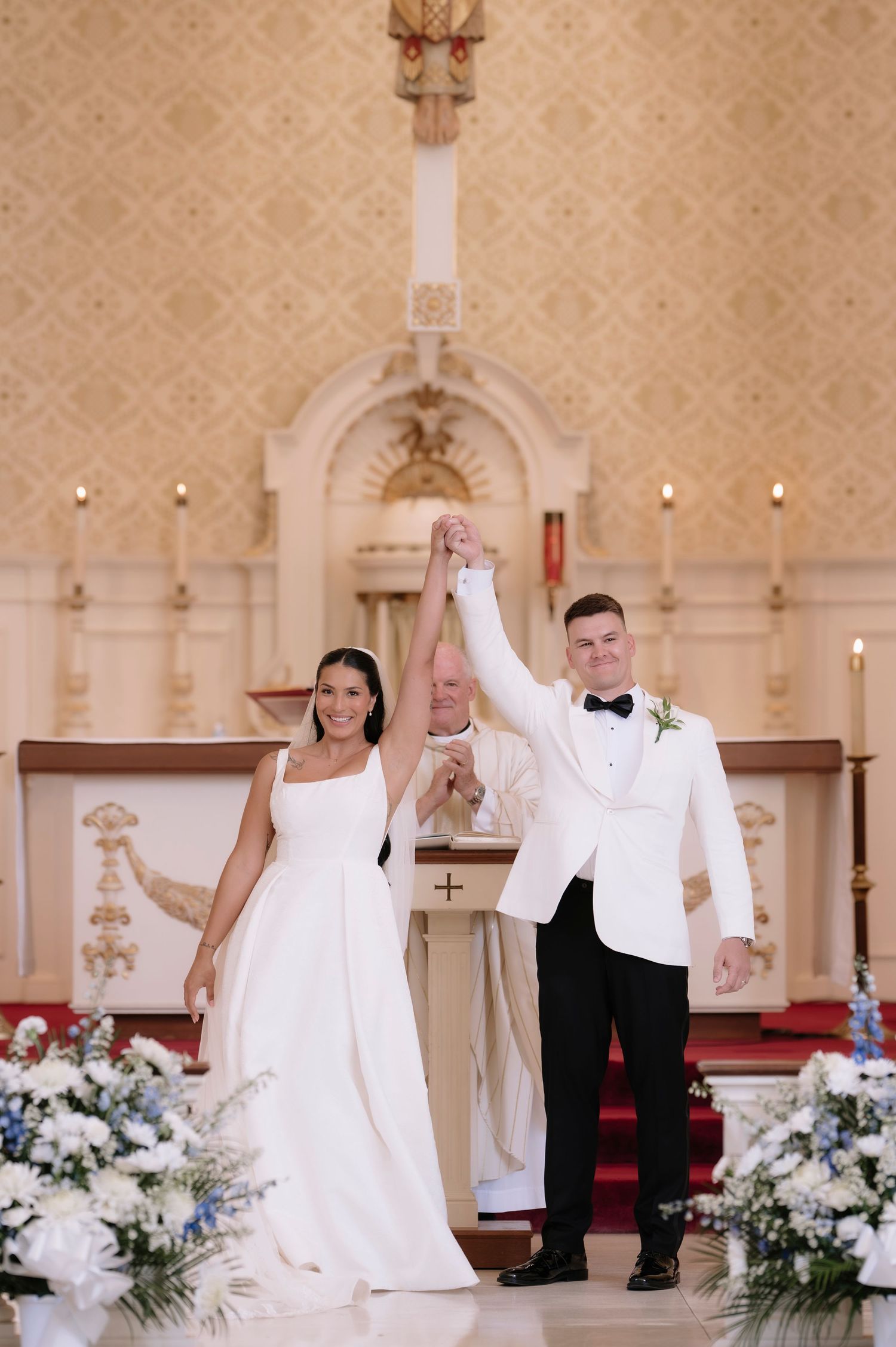 Bride and groom raise hands at church altar after wedding ceremony with floral arrangements.