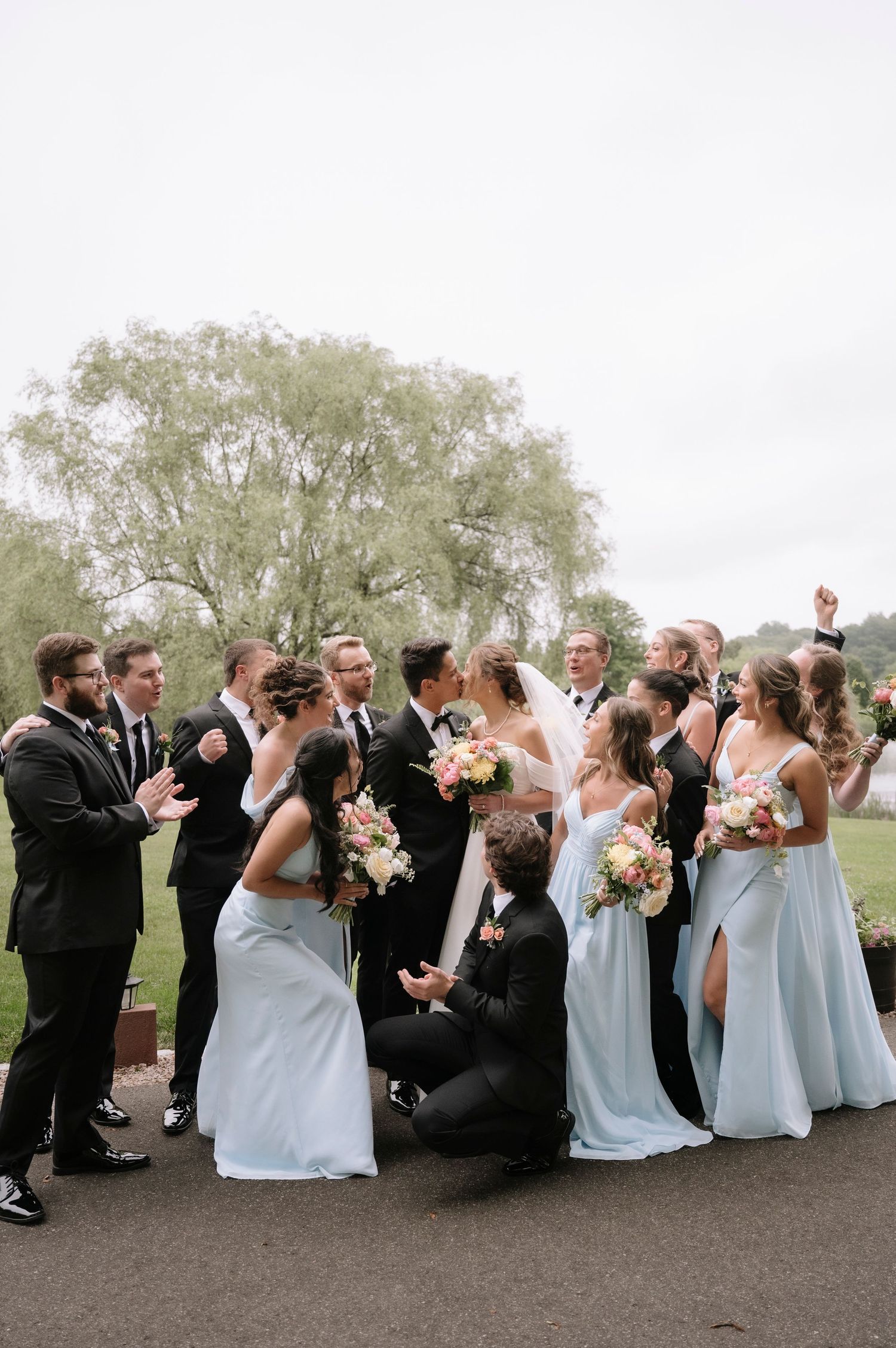 Wedding party poses playfully in light blue dresses and dark suits on asphalt path.