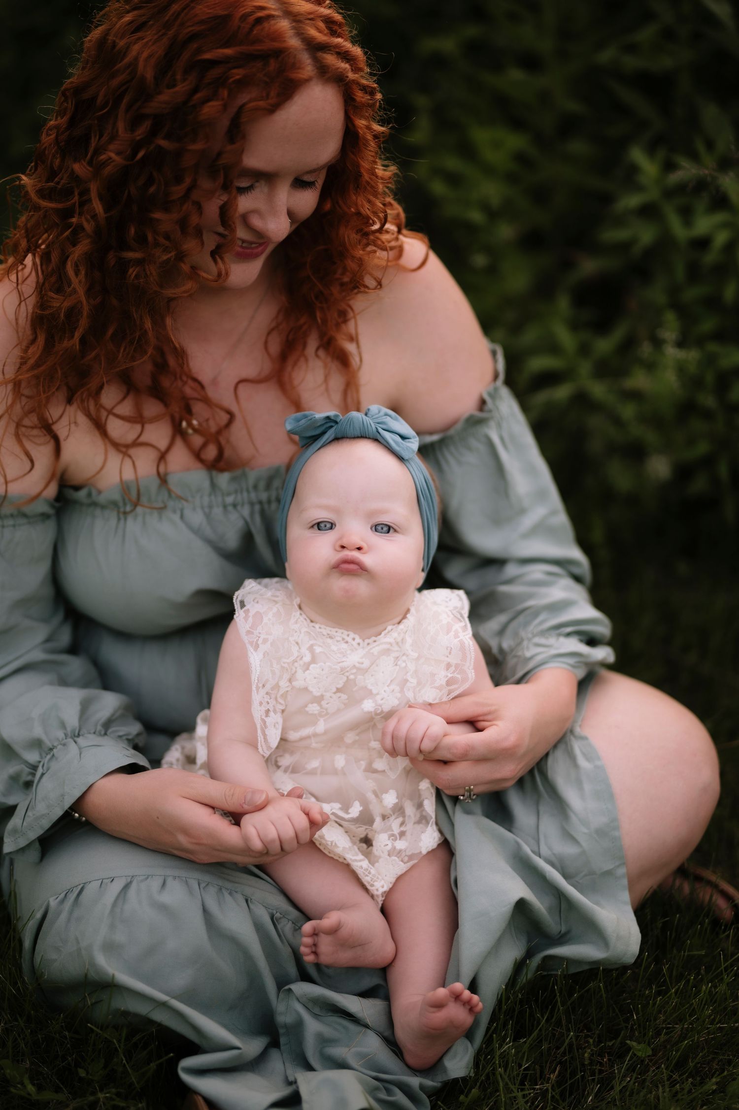 A baby wearing a lace romper and blue headband sits on someone's lap in a grassy outdoor setting.