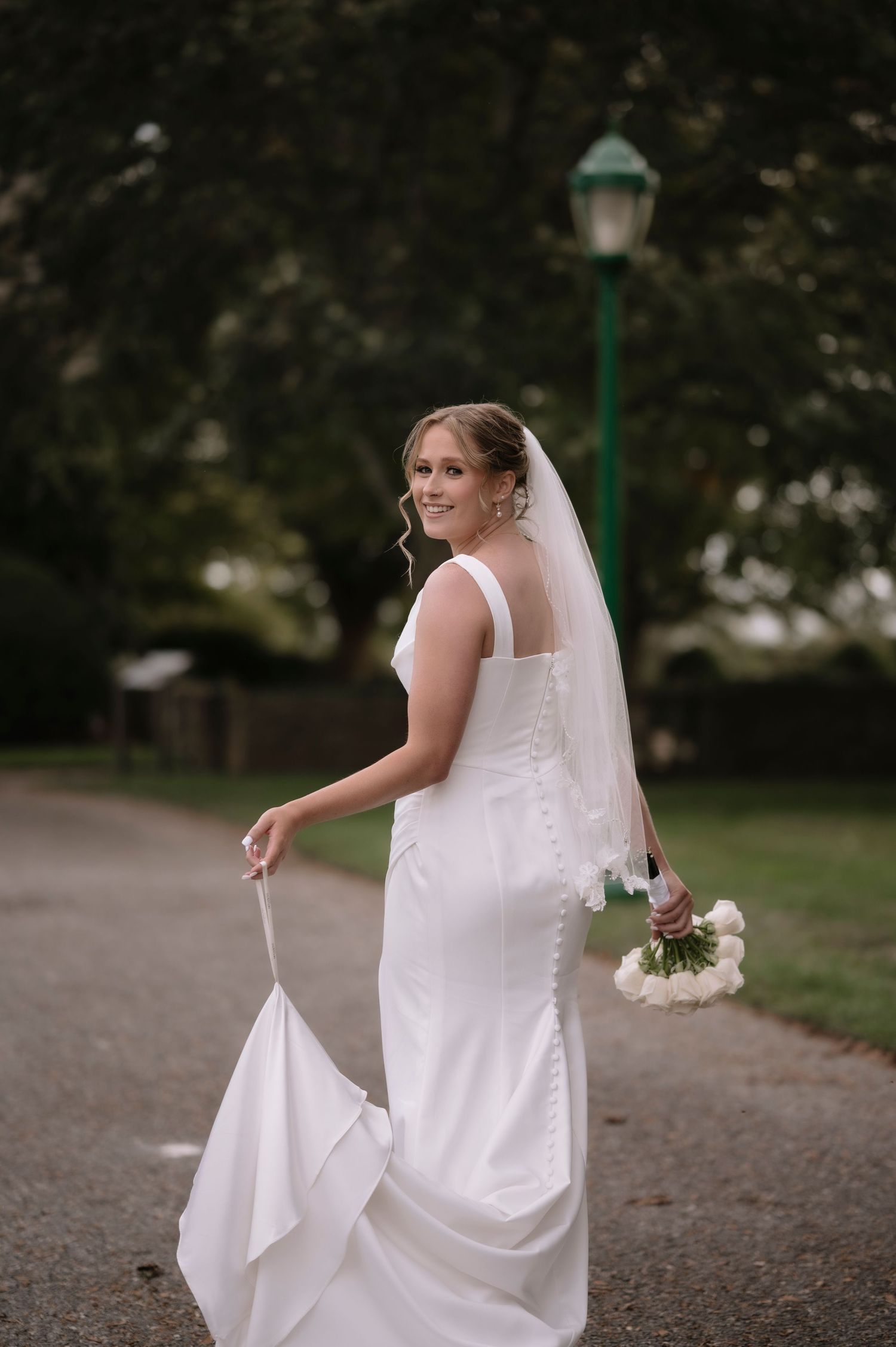 A bride in a white wedding dress holds her bouquet while walking along a path with a green lamppost in the background.