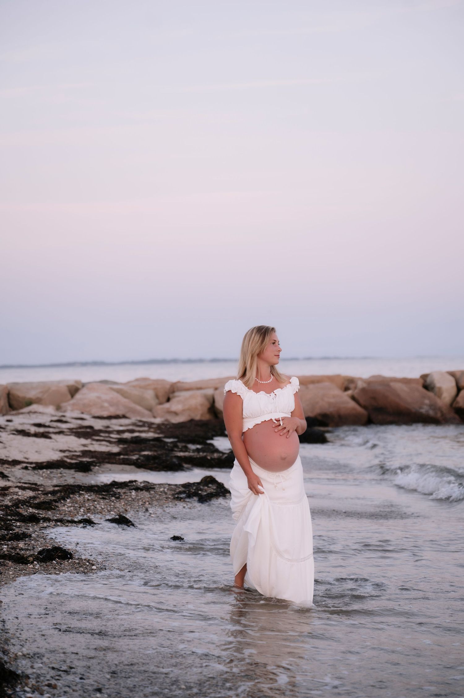 Pregnant woman in white dress standing in ocean waves at beach during golden hour sunset.