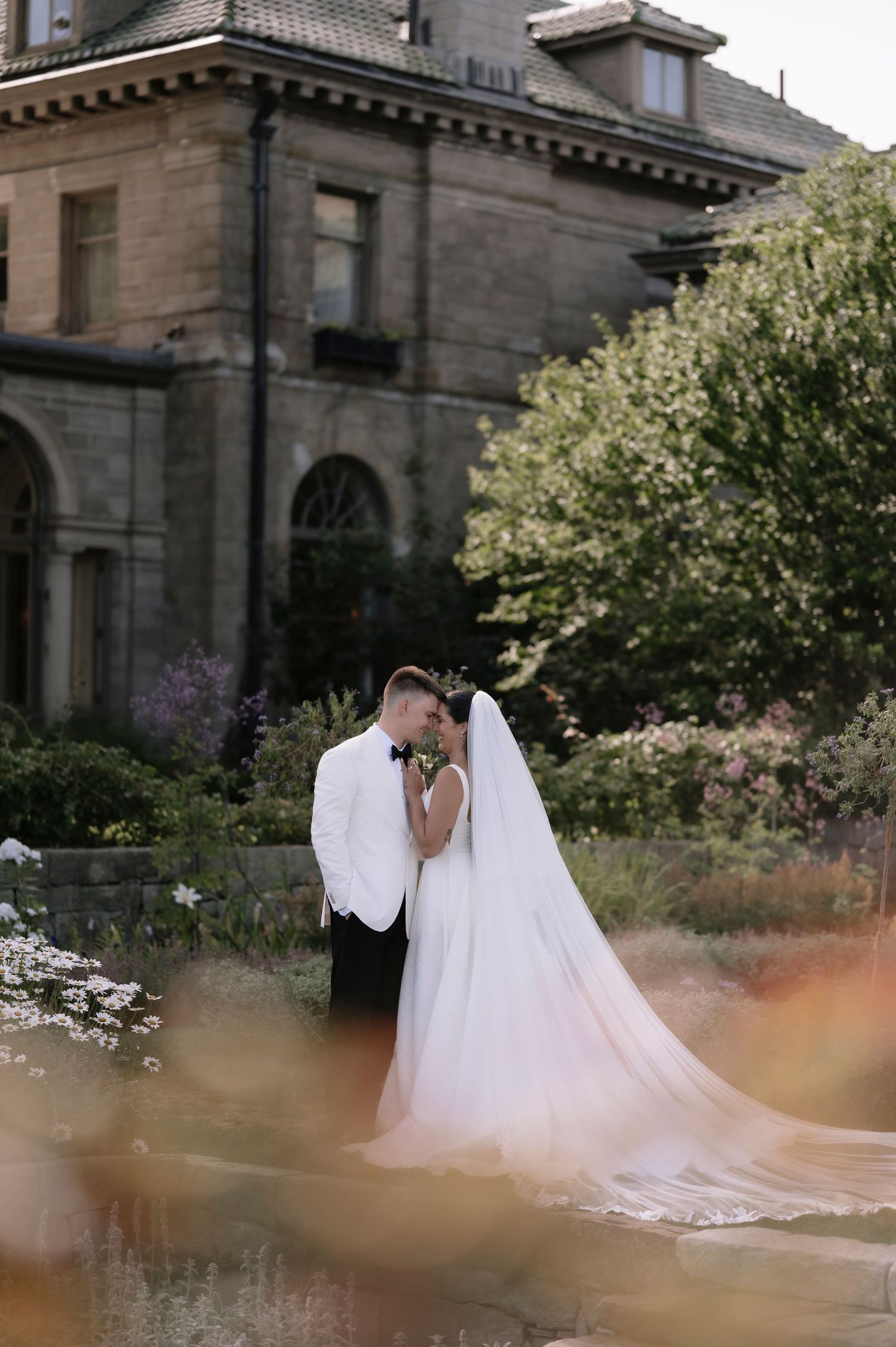 Bride and groom share a romantic kiss in a lush garden outside a grand stone mansion, bride wearing a flowing white gown with veil.