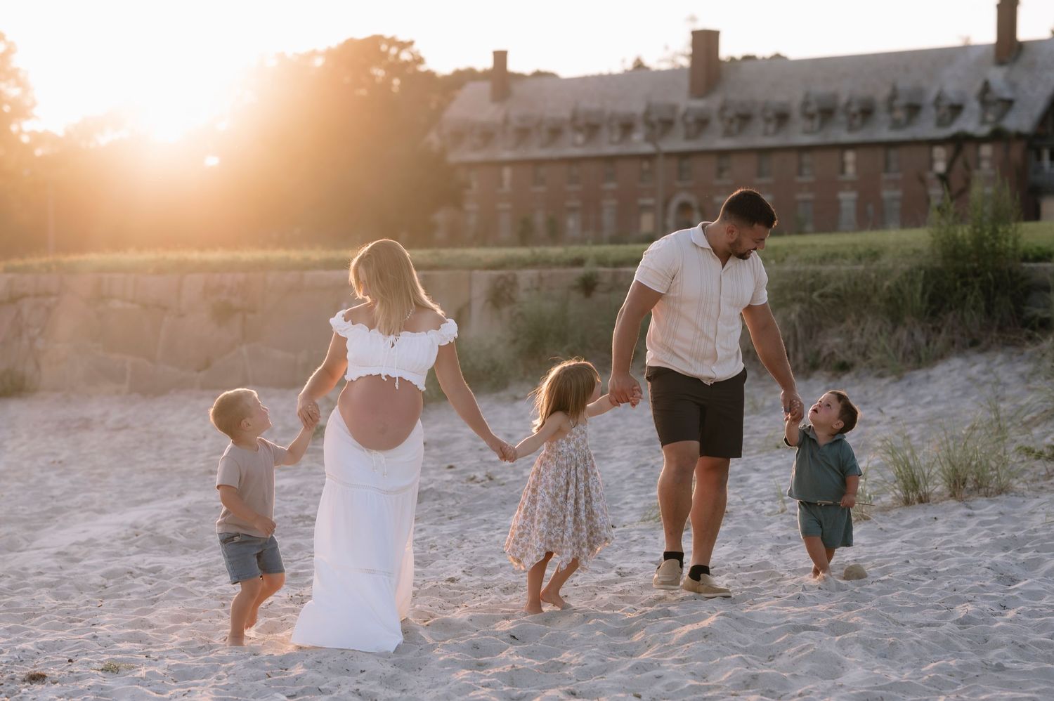 Pregnant woman in white dress with husband and three young children walking on sandy beach at golden sunset.