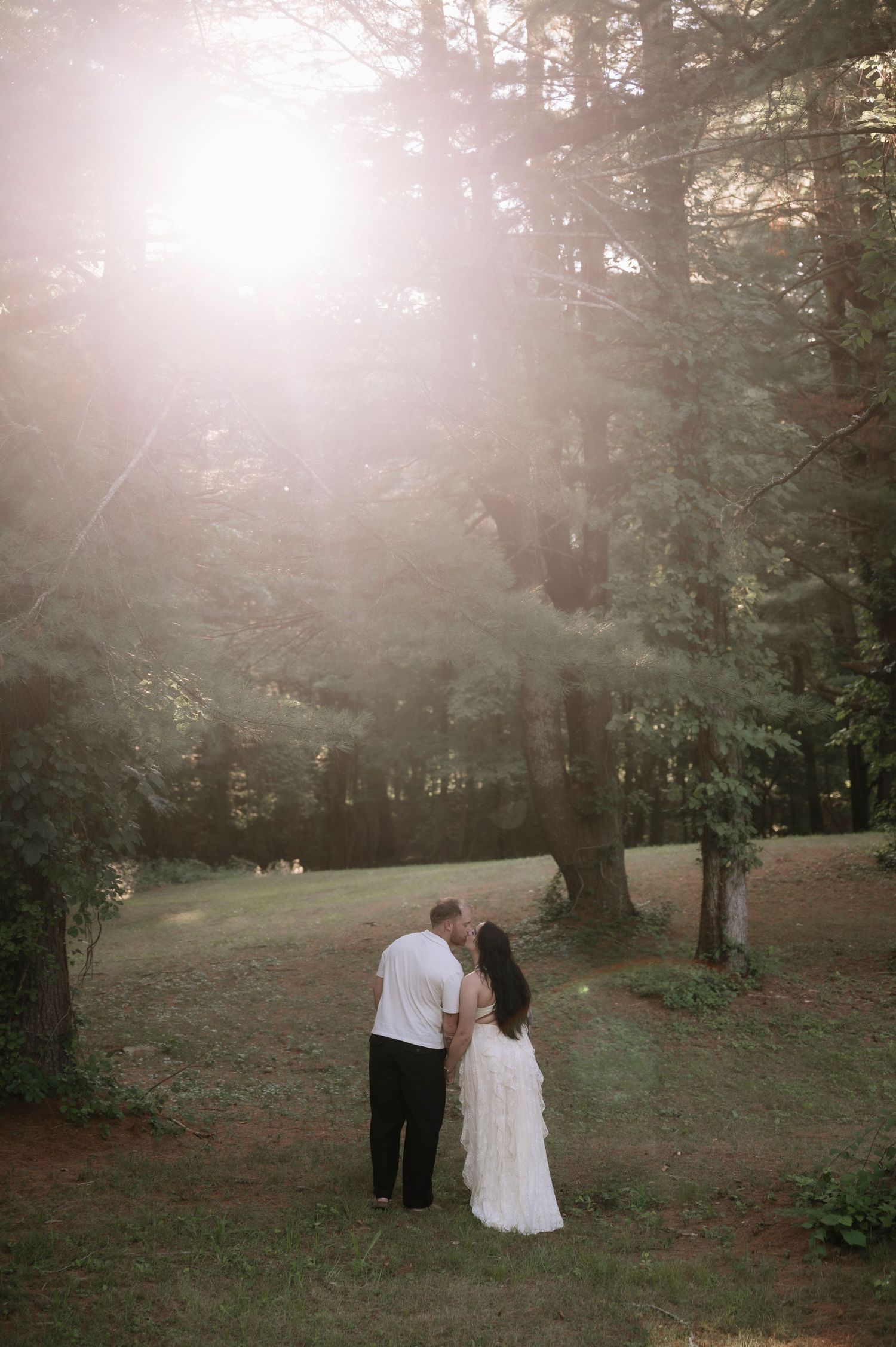 Couple sharing a romantic kiss in a sunlit forest clearing, bride in white gown, groom in black pants and white shirt.