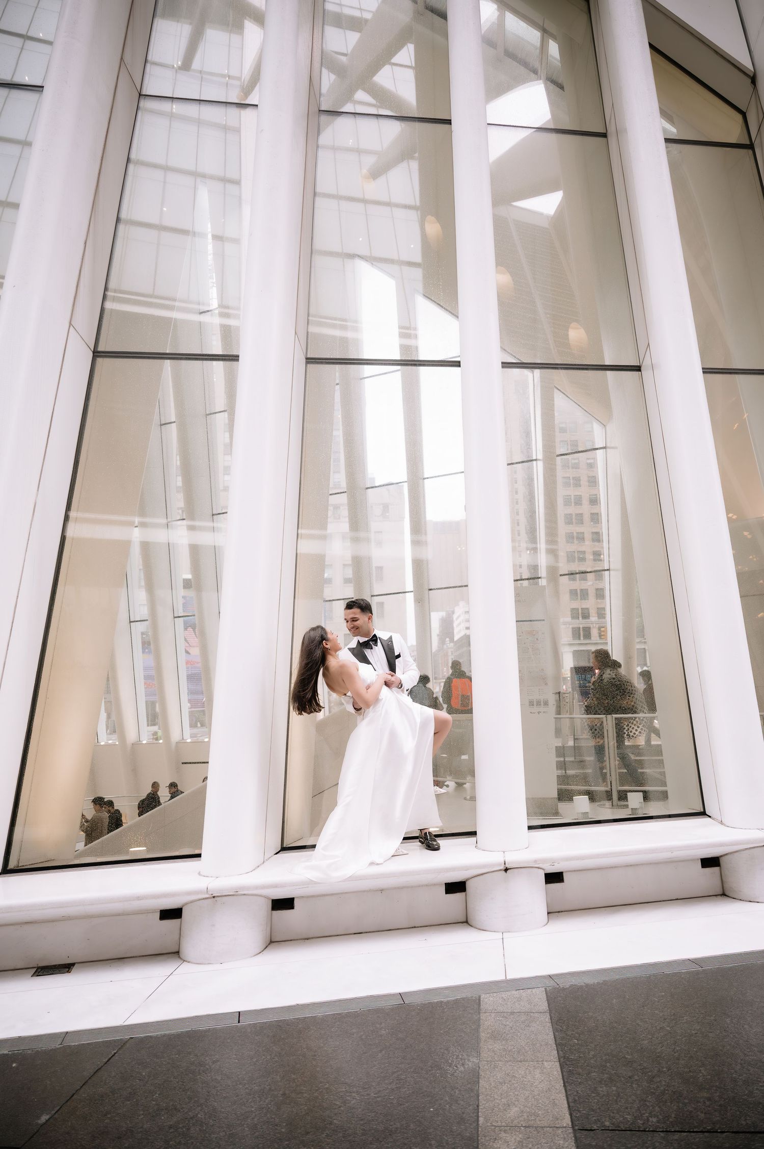 Modern architectural wedding photo with couple posing between white columns and glass windows.