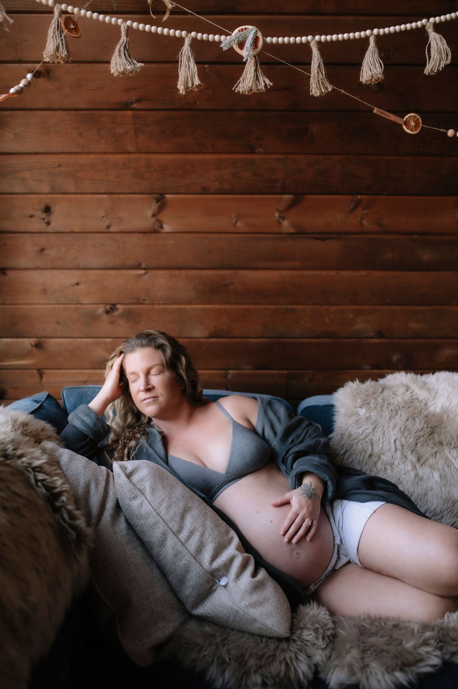 Cozy maternity photo with decorative tassels and fur blankets against rustic wooden wall backdrop.