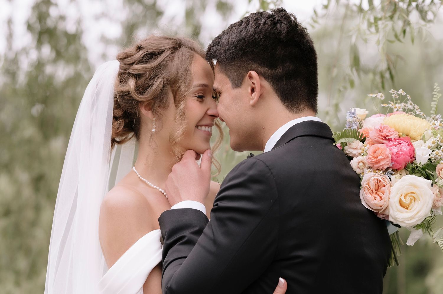 A romantic wedding moment with a bride in white holding pink roses and groom in black suit embracing outdoors.