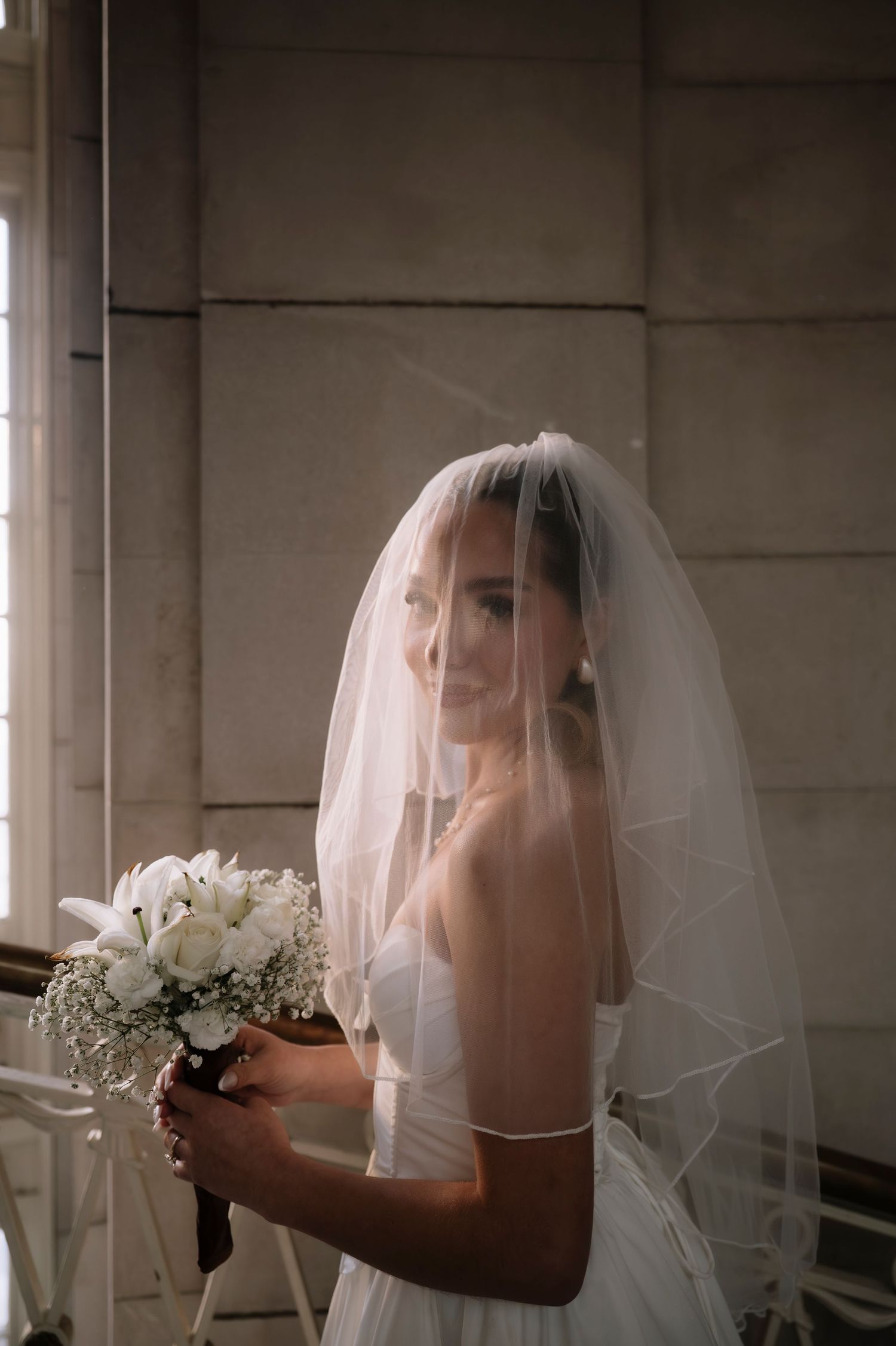 A bride in a white wedding dress and veil holds a bouquet of white flowers in a softly lit interior setting.