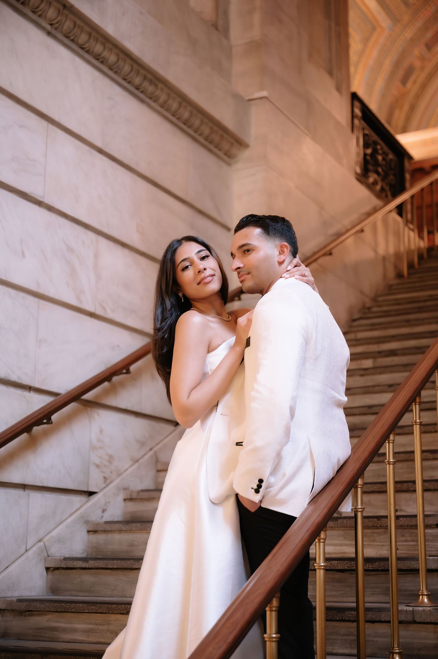 A couple in formal white attire share an intimate moment on an elegant marble staircase with ornate railing.