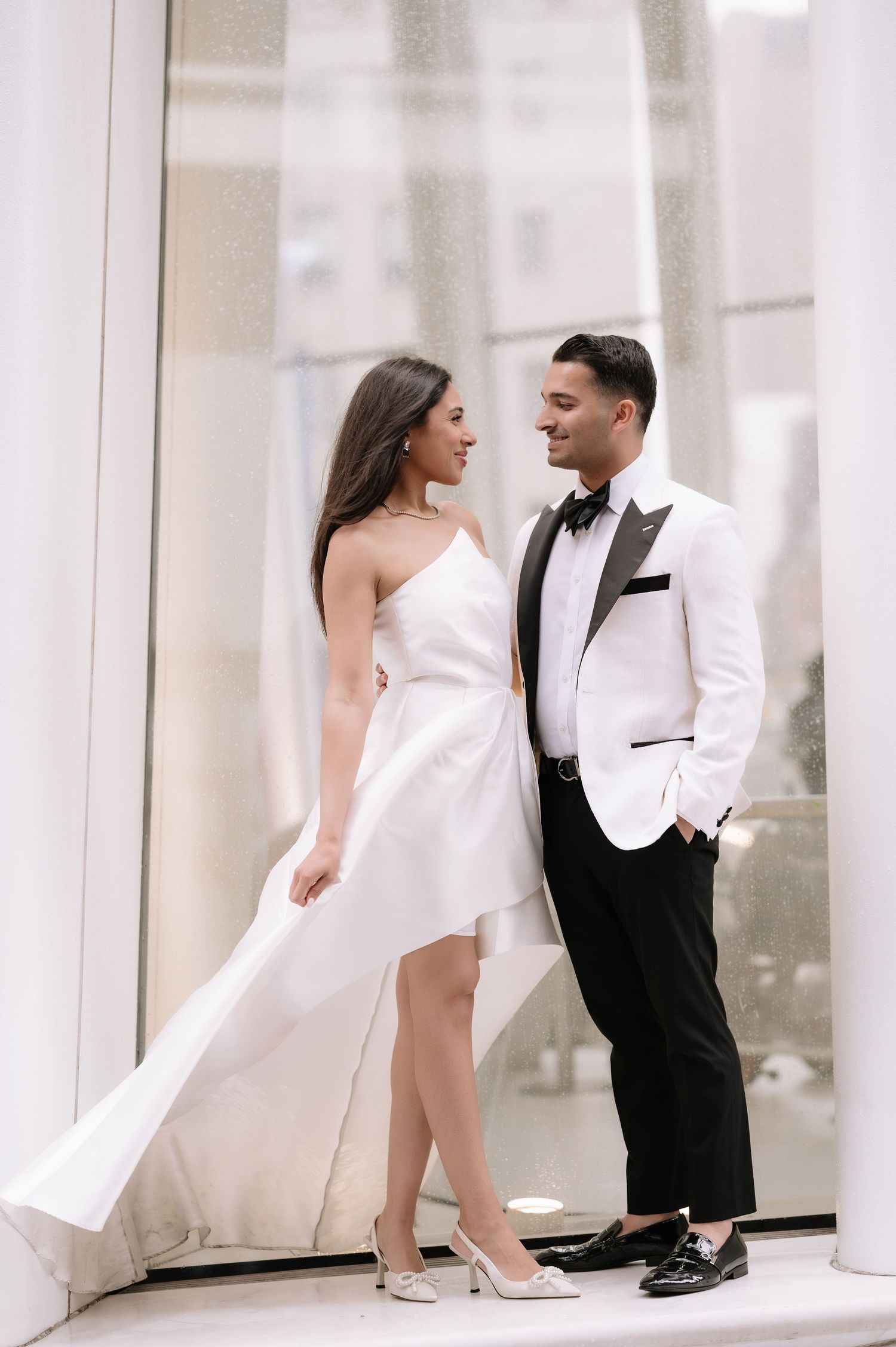Stylish couple in white and black formal wear sharing an intimate moment near modern architecture.