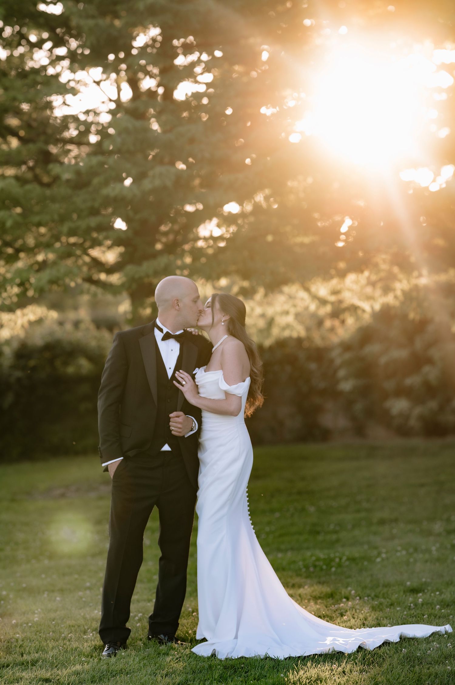 A couple shares a romantic kiss on their wedding day during golden hour sunset in a grassy outdoor setting.