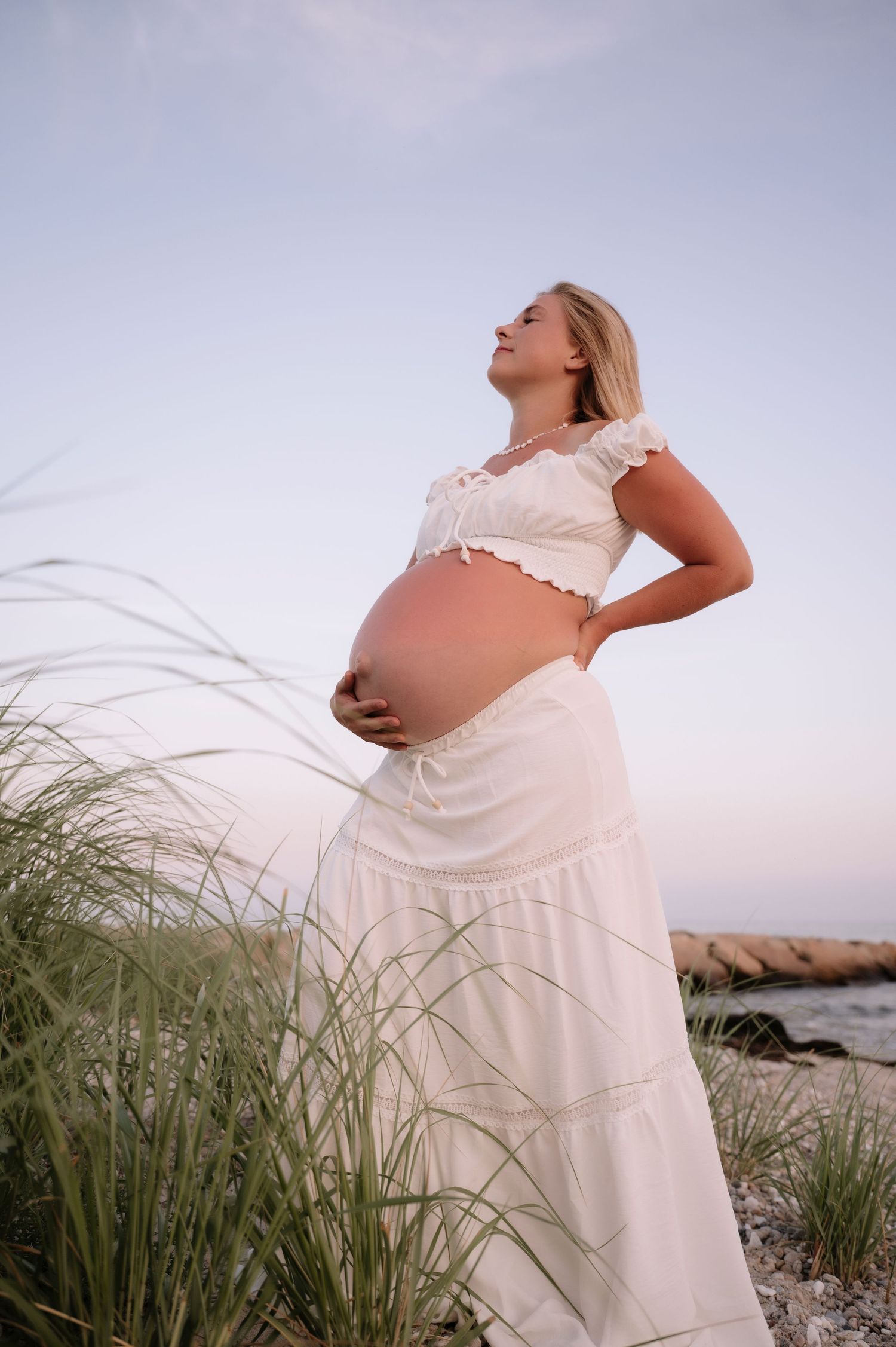 Pregnant woman in white crop top and maxi skirt holding belly on beach at sunset.
