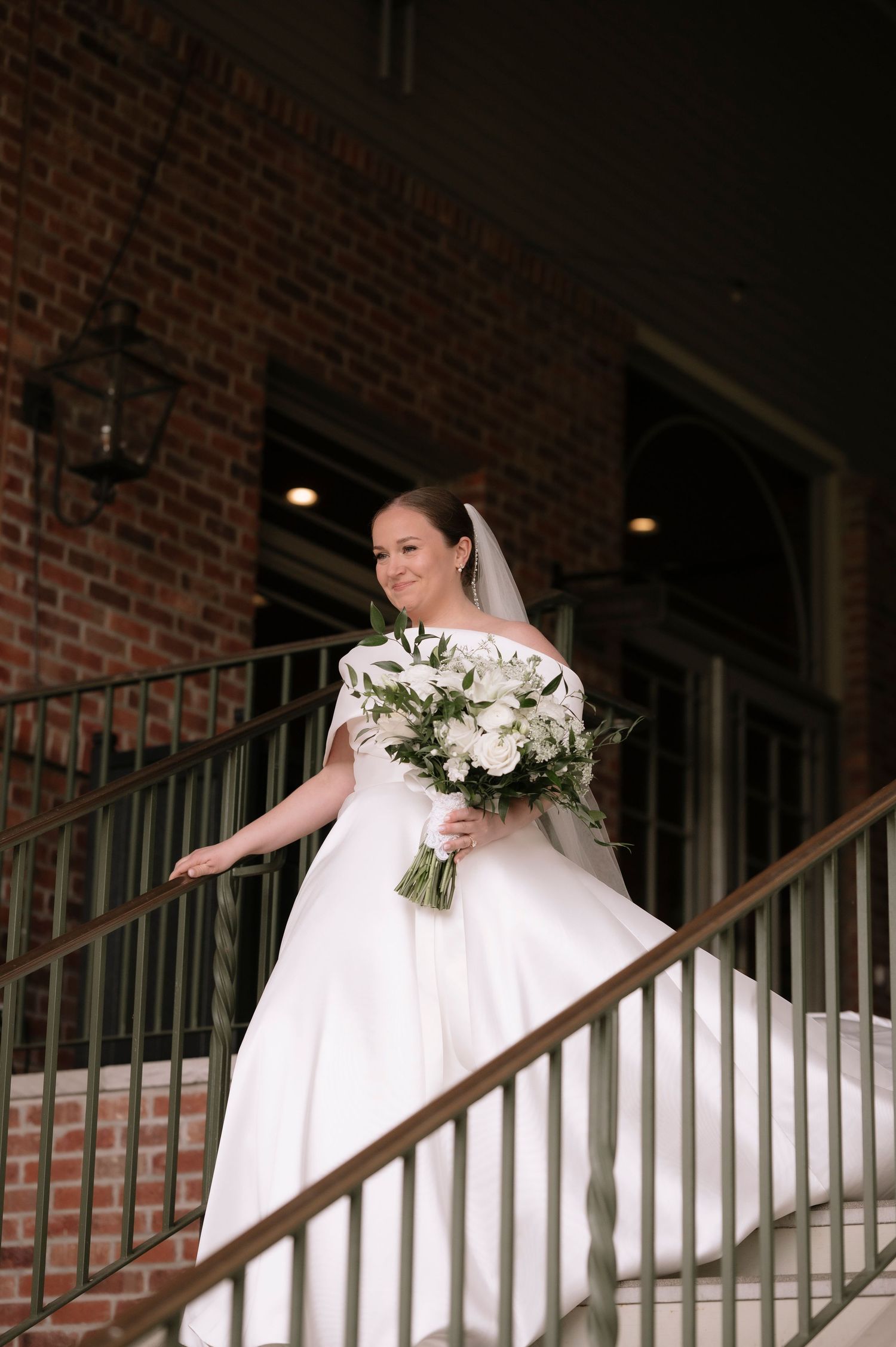 Bride in white off-shoulder gown holds floral bouquet on brick building staircase, veil flowing behind her.