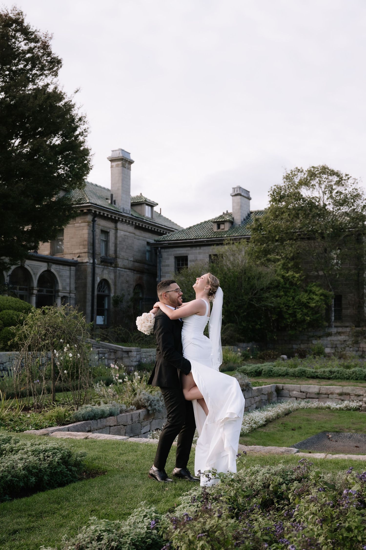 A couple shares romantic moments in a garden setting with historic stone buildings in the background.