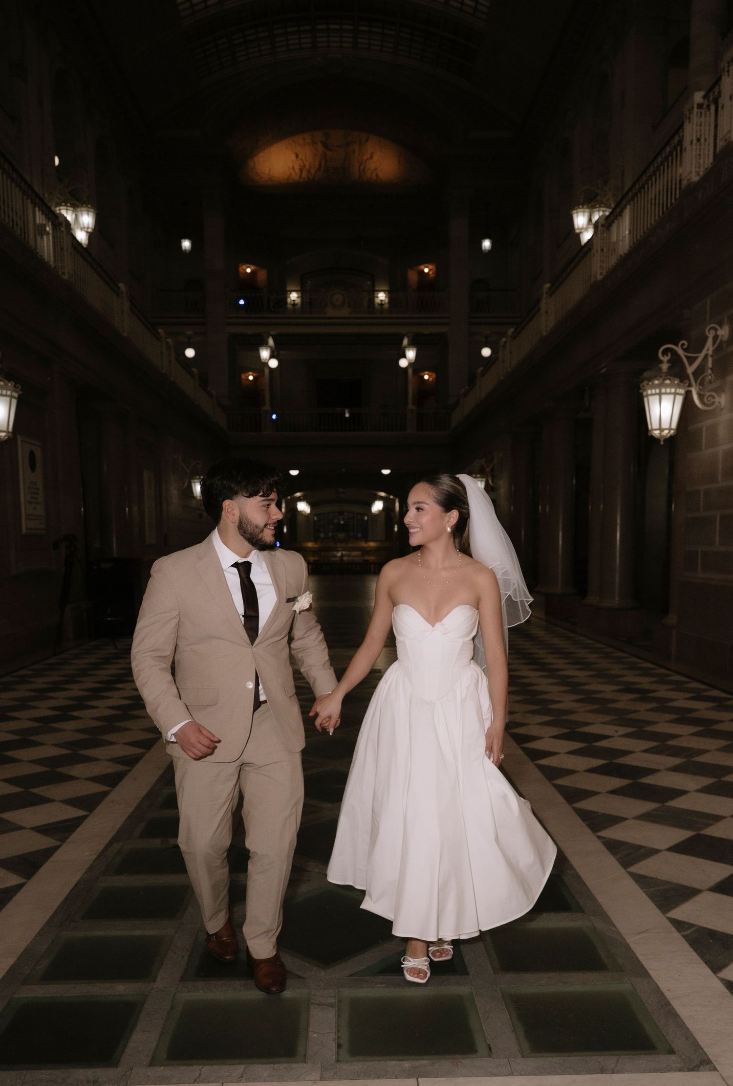 A couple in wedding attire walks across a checkered floor in a dimly lit venue at night.