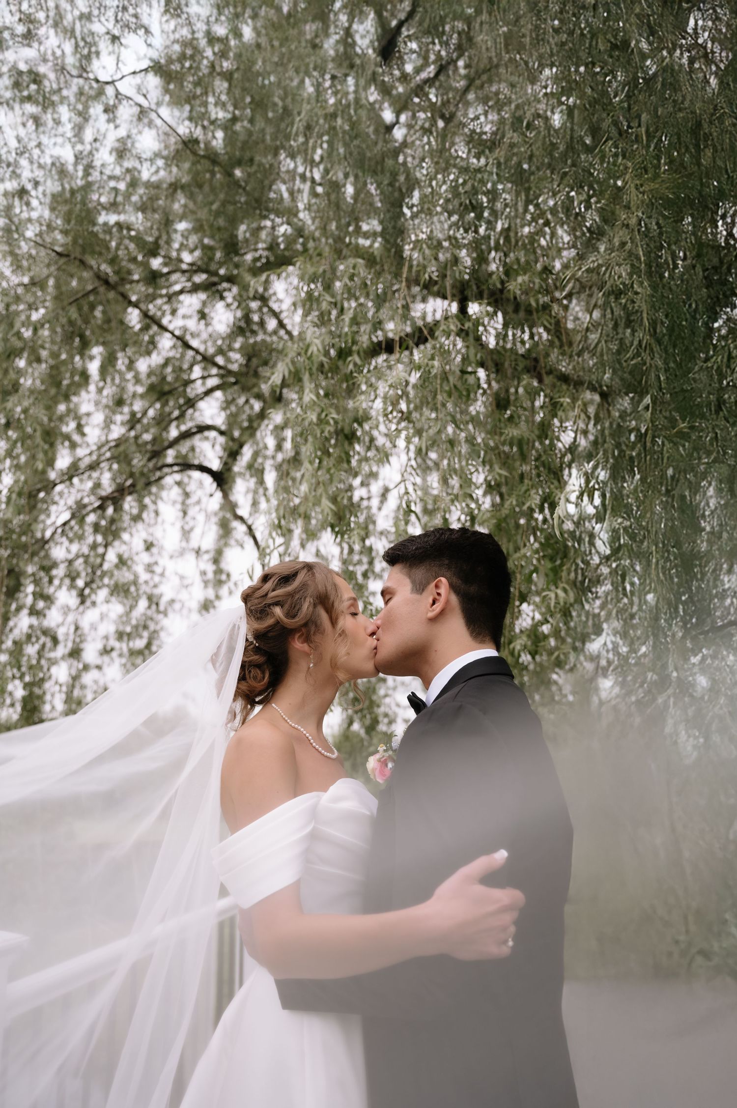 Romantic wedding couple sharing intimate moments under a willow tree with flowing veil.
