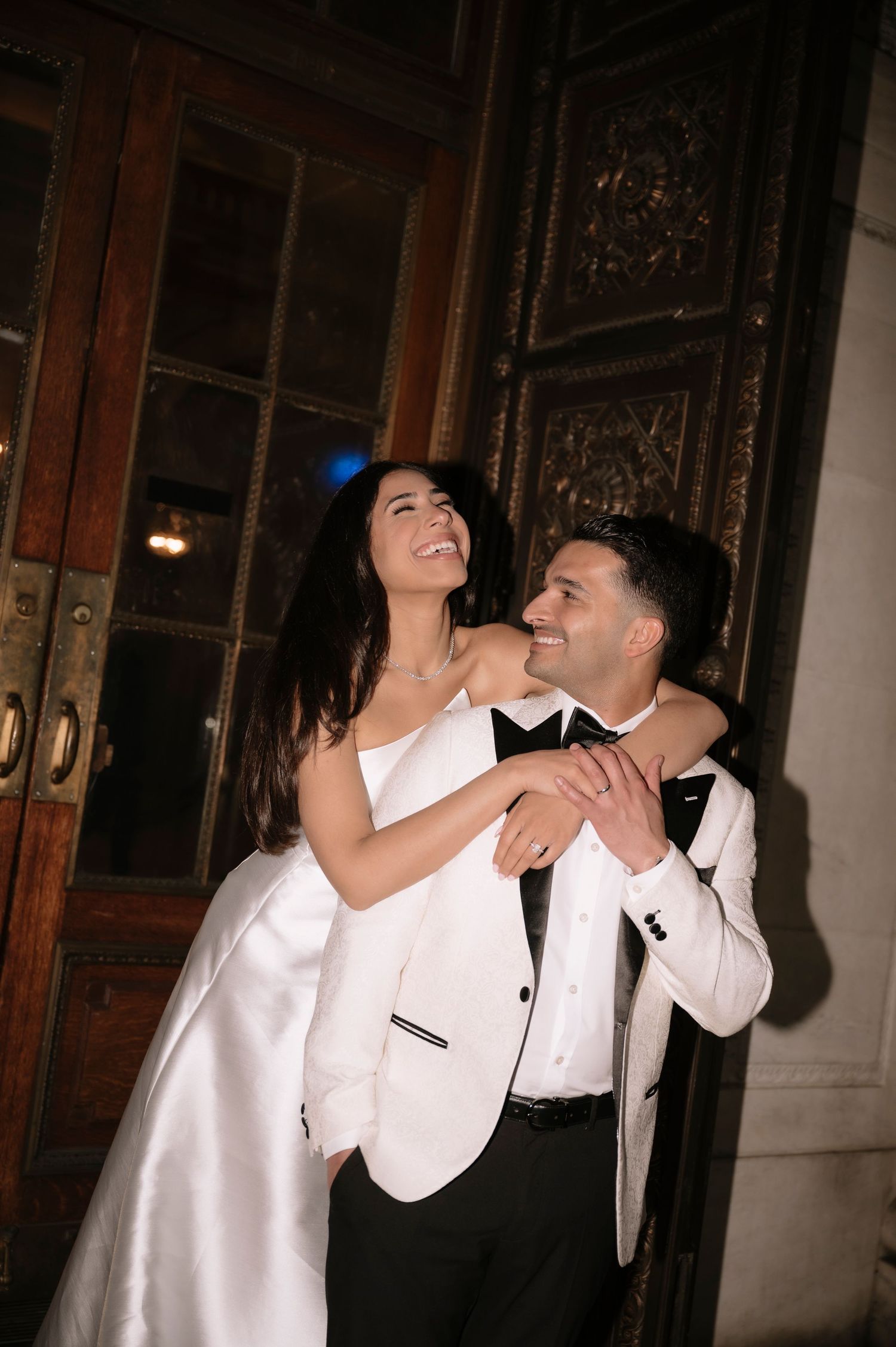 A joyful couple shares a candid moment together at their wedding, dressed in white formal attire by elegant wooden doors.