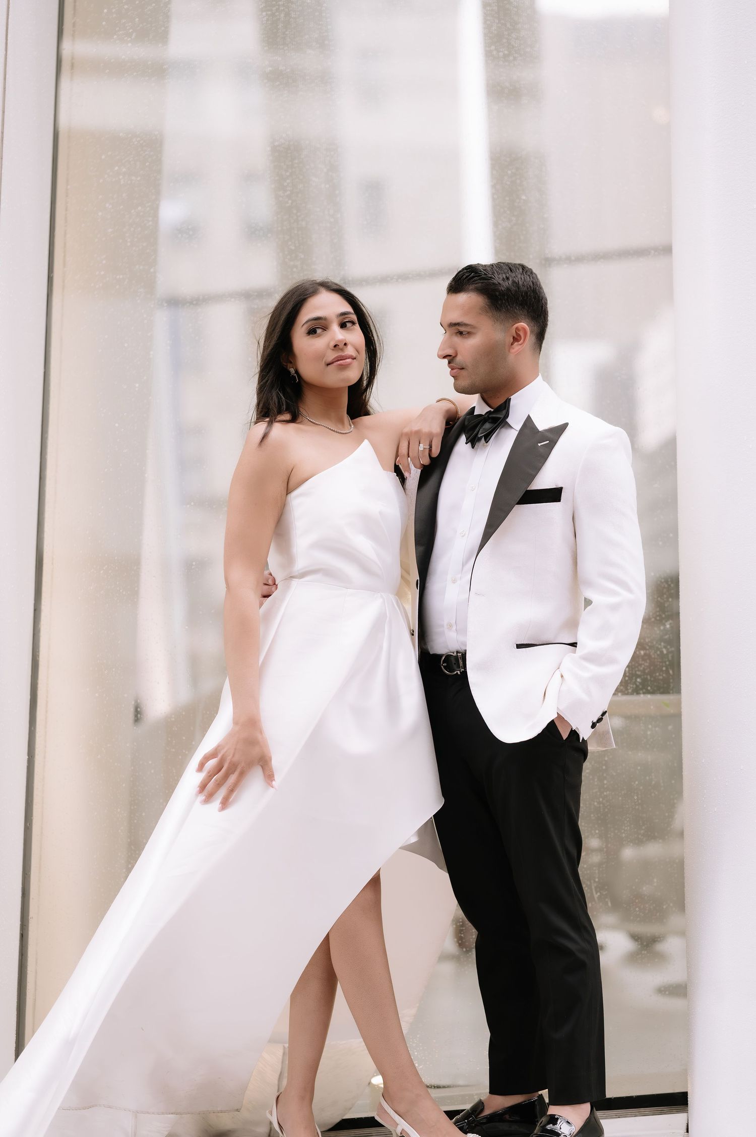 Elegant couple in formal attire posing against white columns, bride wearing a flowing white gown with slit.